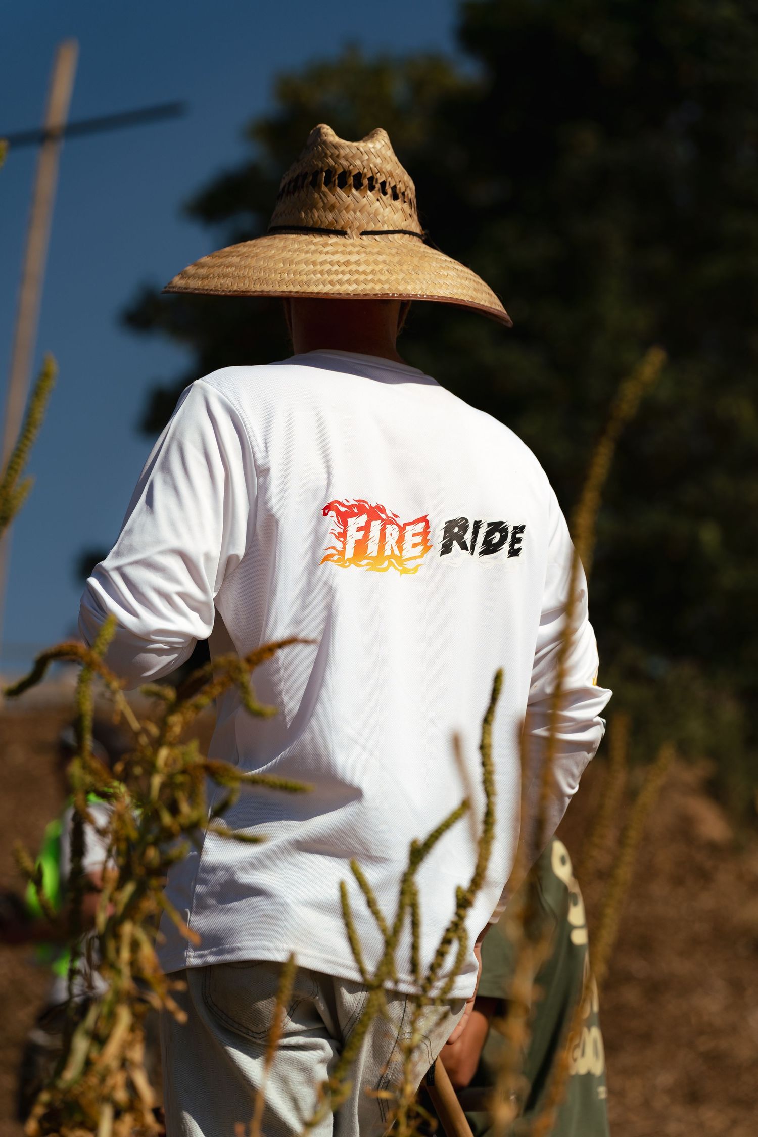 Man wearing straw hat and white Fire Ride long sleeve shirt standing outdoors among desert plants.