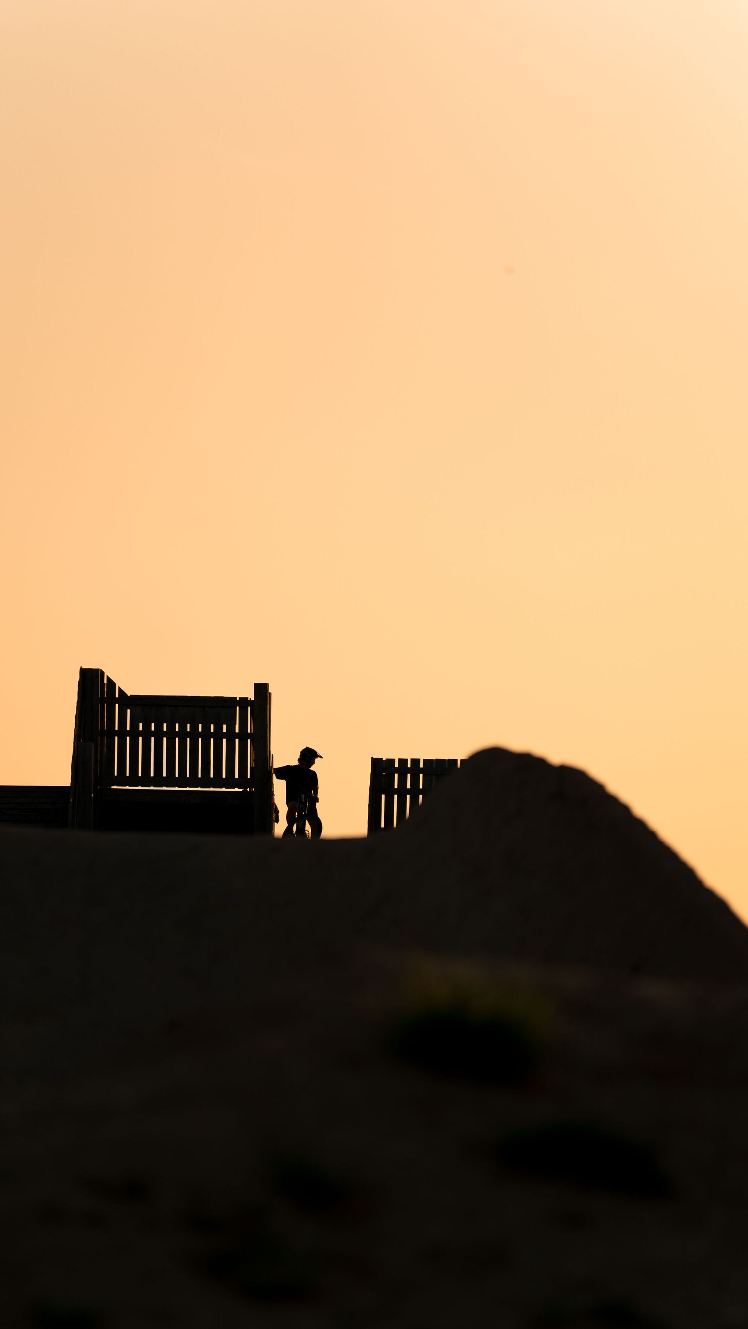 Silhouette of a person standing near wooden fence gates on a hill against a warm golden sunset sky.