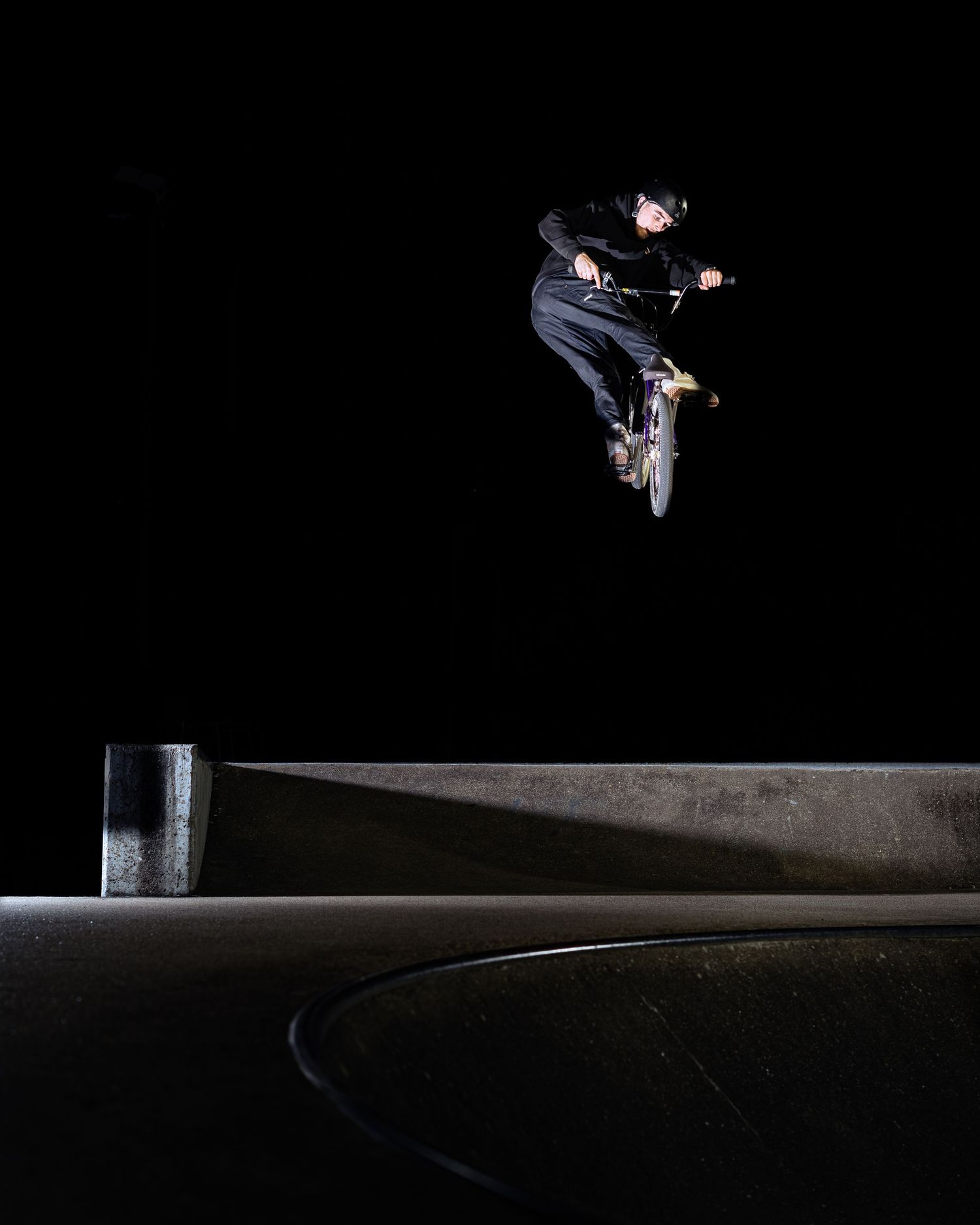 BMX rider performing a high aerial trick at night over a concrete skate park ramp.