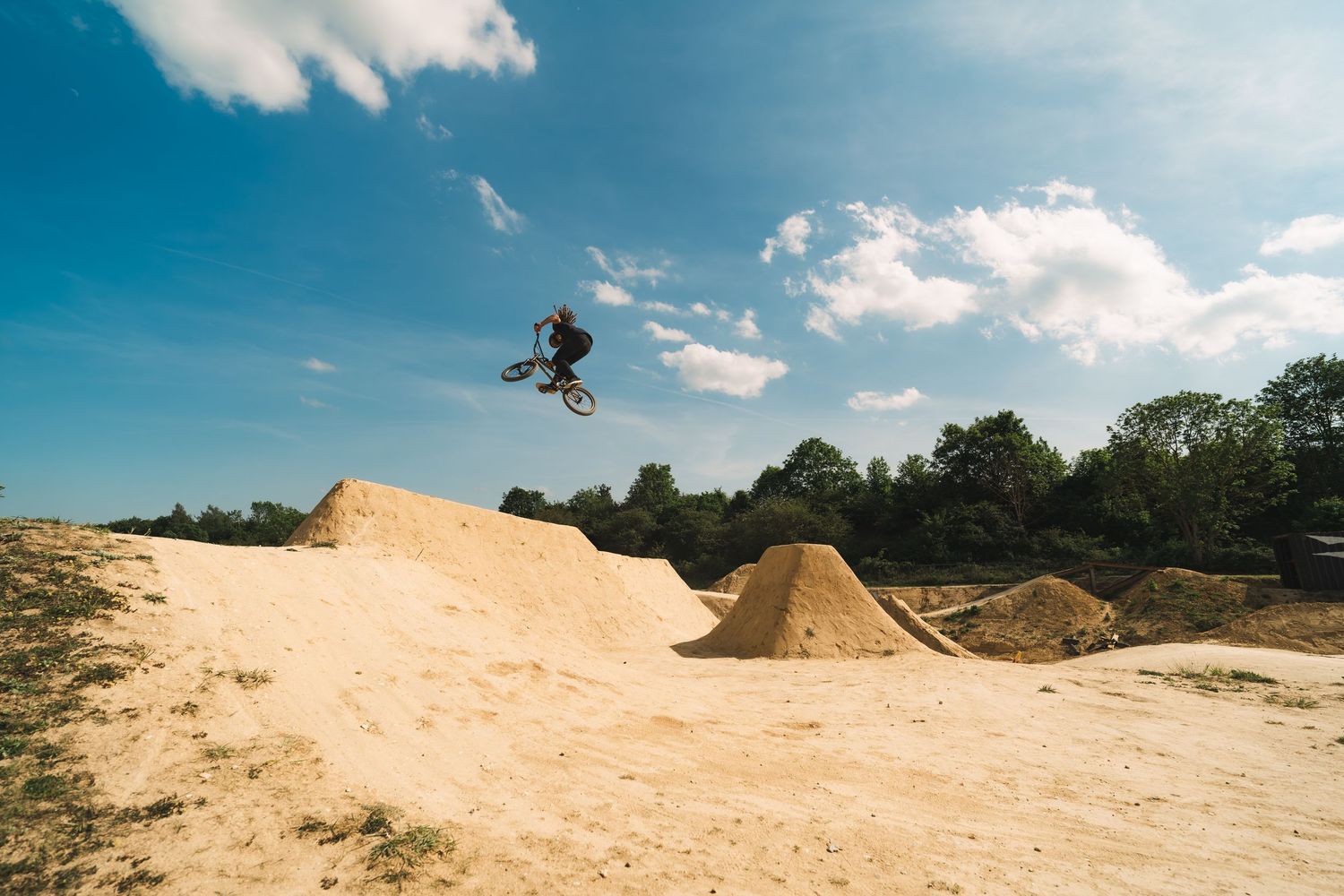 BMX rider catching air over a series of sandy dirt jumps at an outdoor riding park with trees in background.