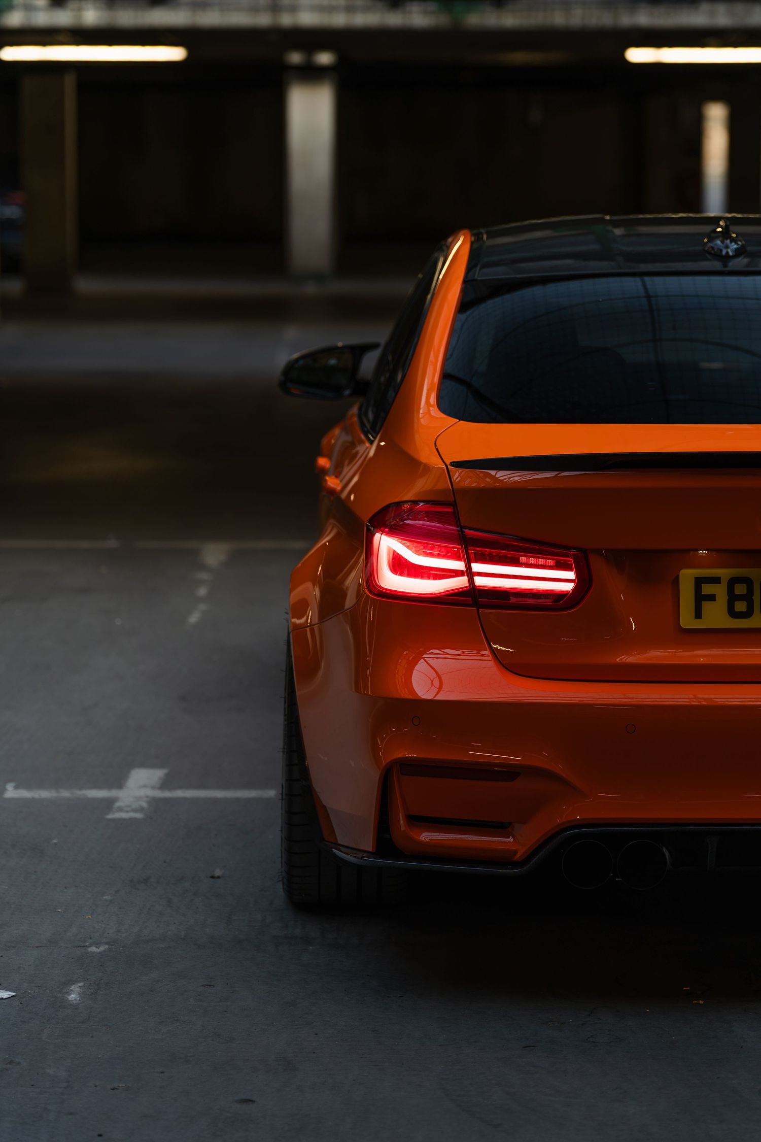 Rear view of a vibrant orange BMW M2 with glowing red taillights parked in a dimly lit parking garage.