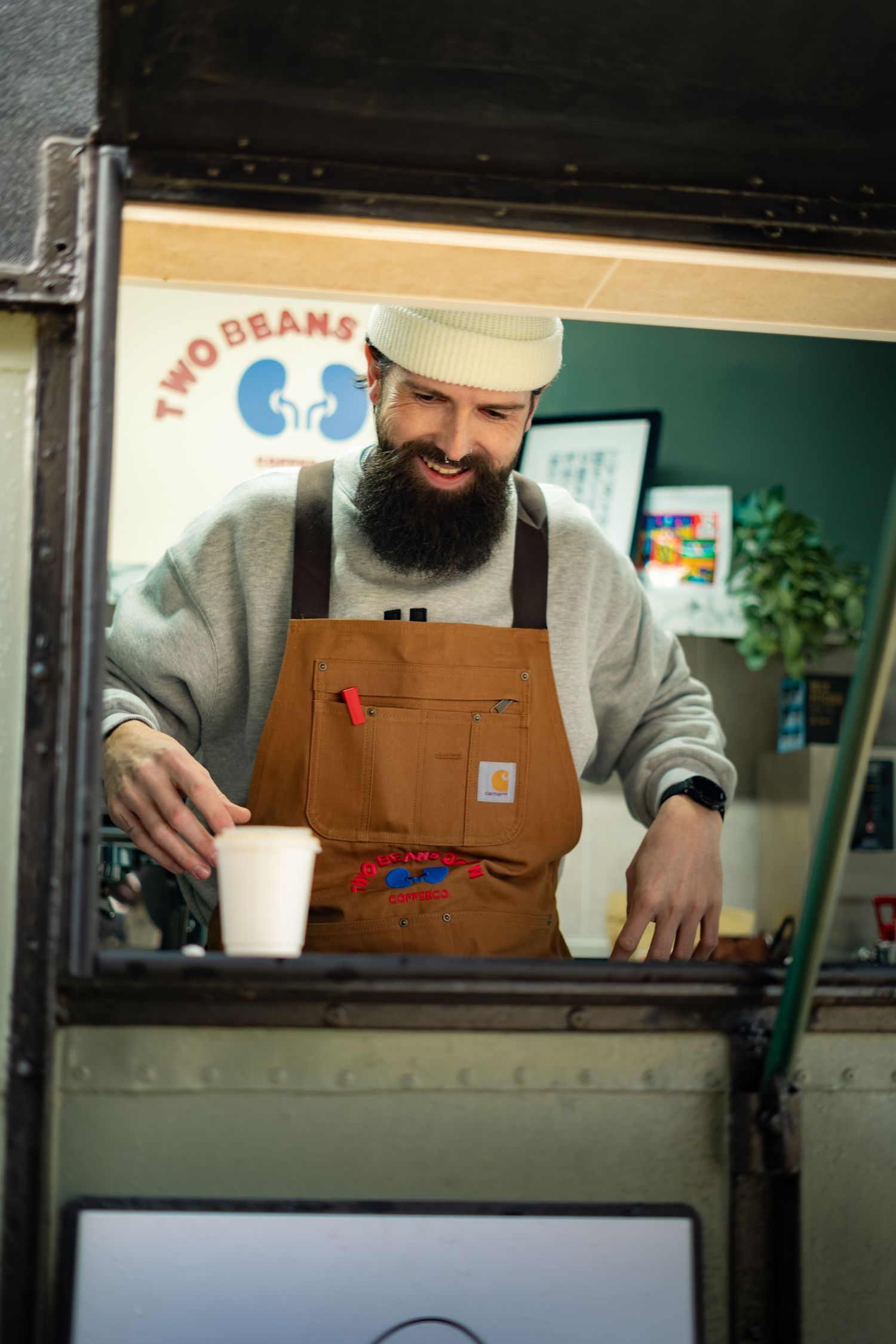 Smiling bearded barista serving coffee inside Two Beans Down Coffee Co. mobile trailer.