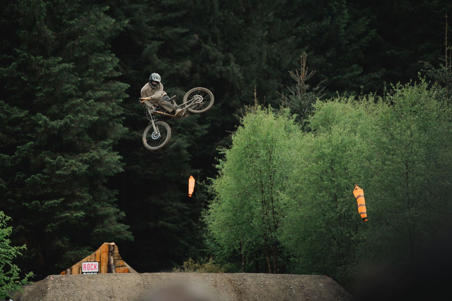 A mountain biker performs a high aerial jump trick over a dirt ramp surrounded by lush green forest trees.