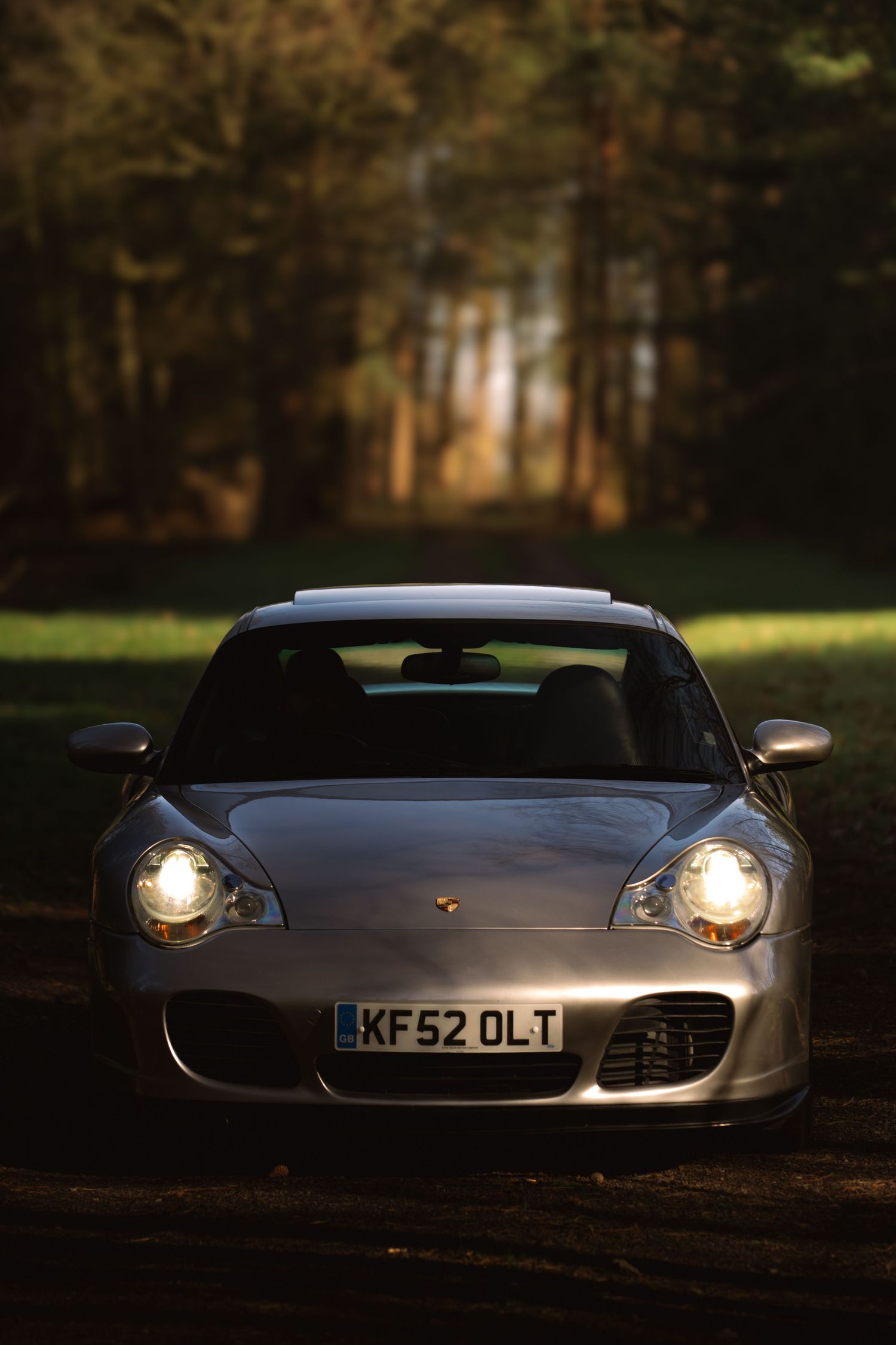 Front view of a silver Porsche 911 Turbo with UK plate KF52 OLT lit by headlights in a forest.