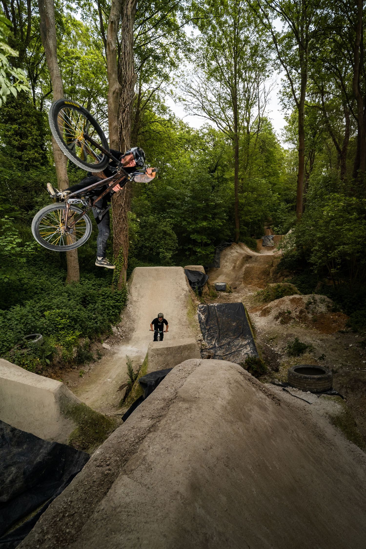 BMX rider performs an aerial trick at a forest dirt jump park while another cyclist watches below.