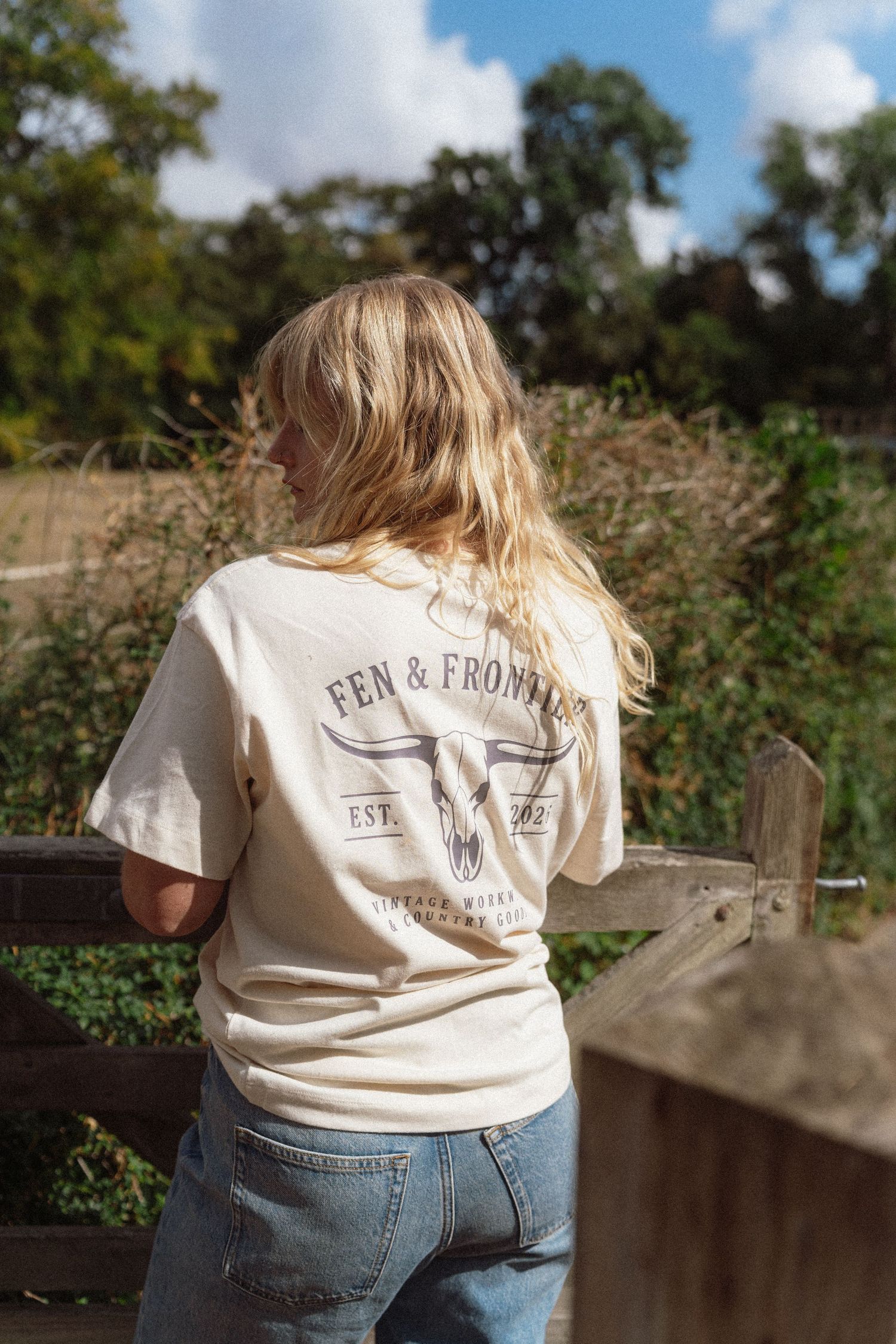 Woman wearing a cream Fen & Frontier EST. 2022 graphic t-shirt with jeans, standing by a wooden fence outdoors.
