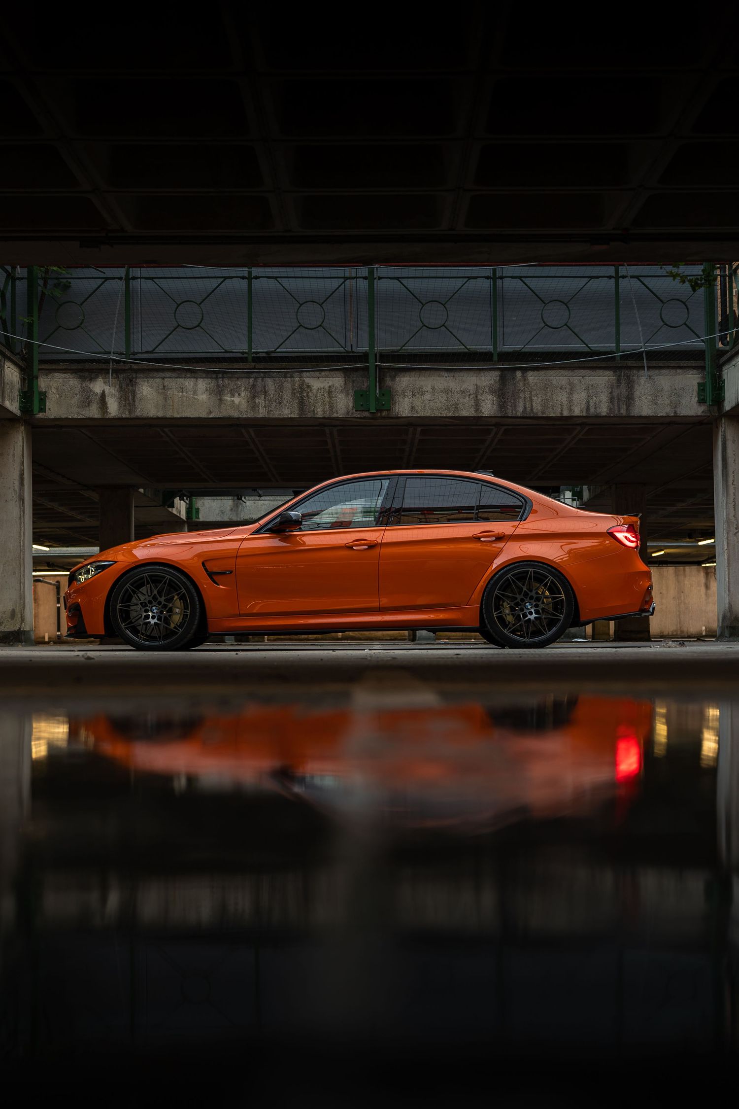 Orange BMW M3 parked in a dark garage with a vivid reflection on the wet floor below.