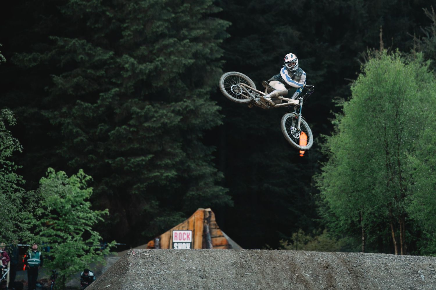 A mountain biker performs a high aerial jump trick over a dirt ramp surrounded by dense green forest trees.