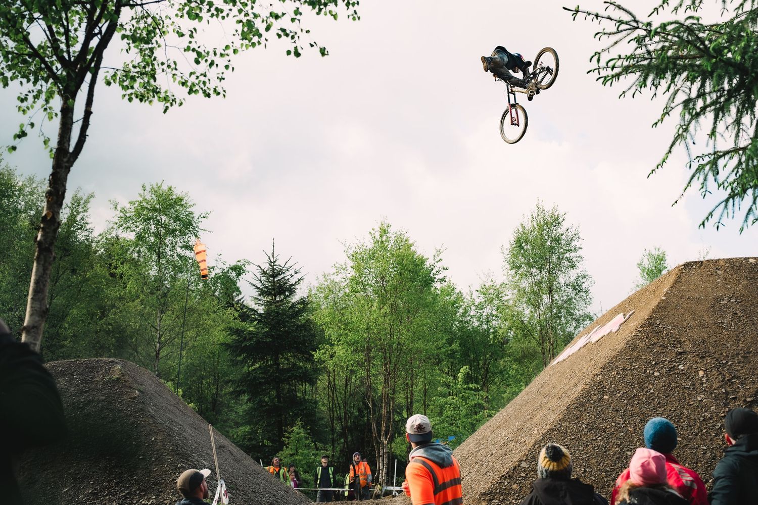 A mountain biker performs a high aerial trick over dirt jumps at a bike park, with spectators watching and trees in the background.