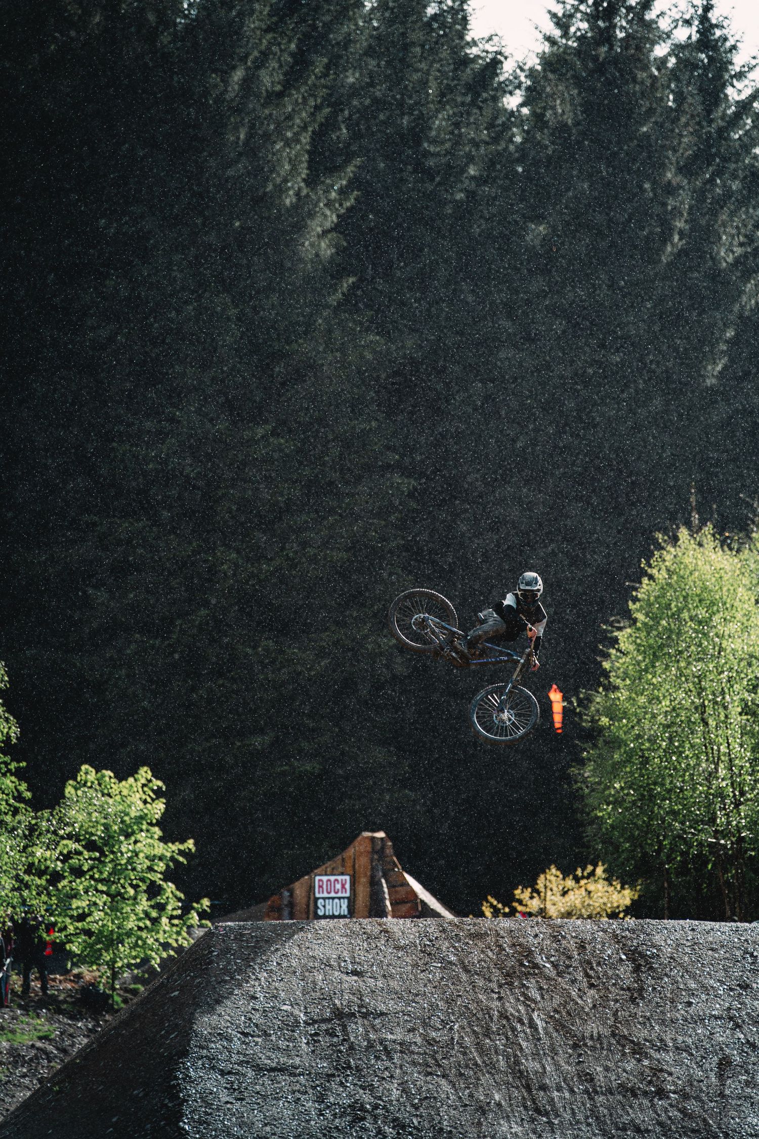 A mountain biker performs a daring aerial trick off a dirt jump ramp surrounded by dense evergreen forest.