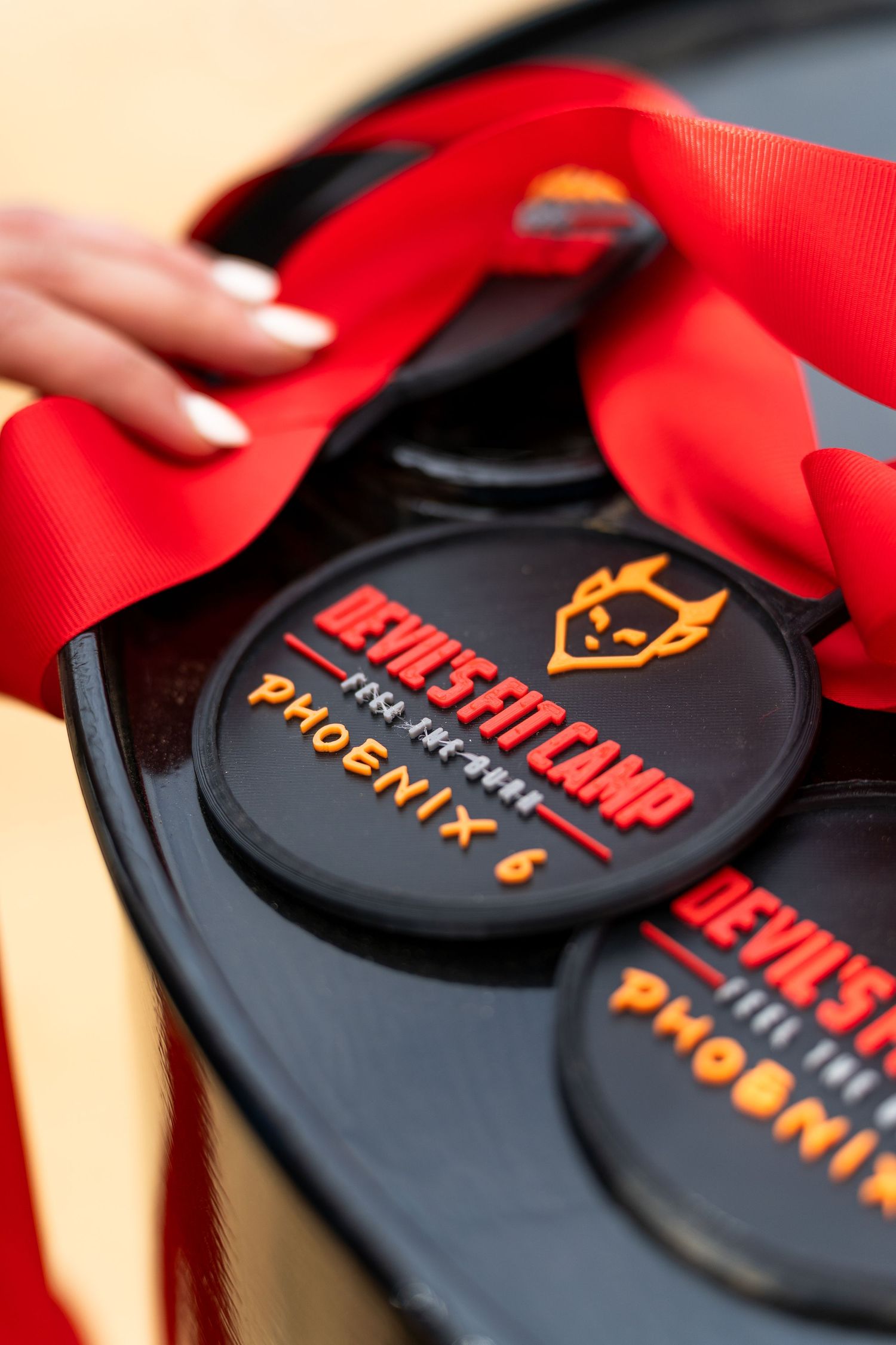 Close-up of Devil's Fit Camp Phoenix 6 medals with red ribbon, held by a woman with white manicured nails.