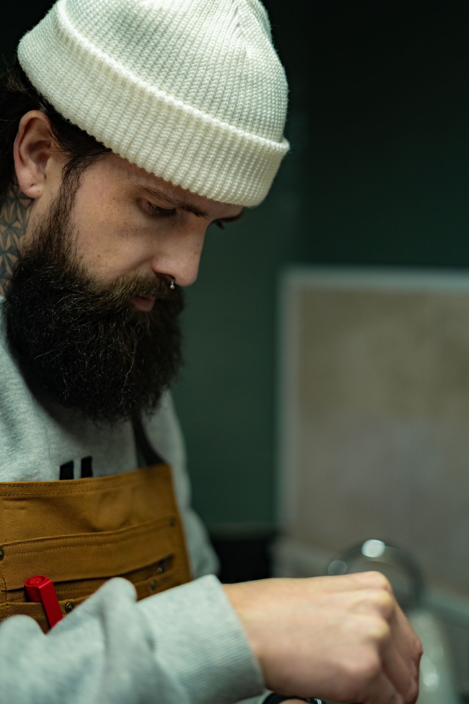Focused bearded barista in white beanie and brown apron preparing coffee at an espresso machine.