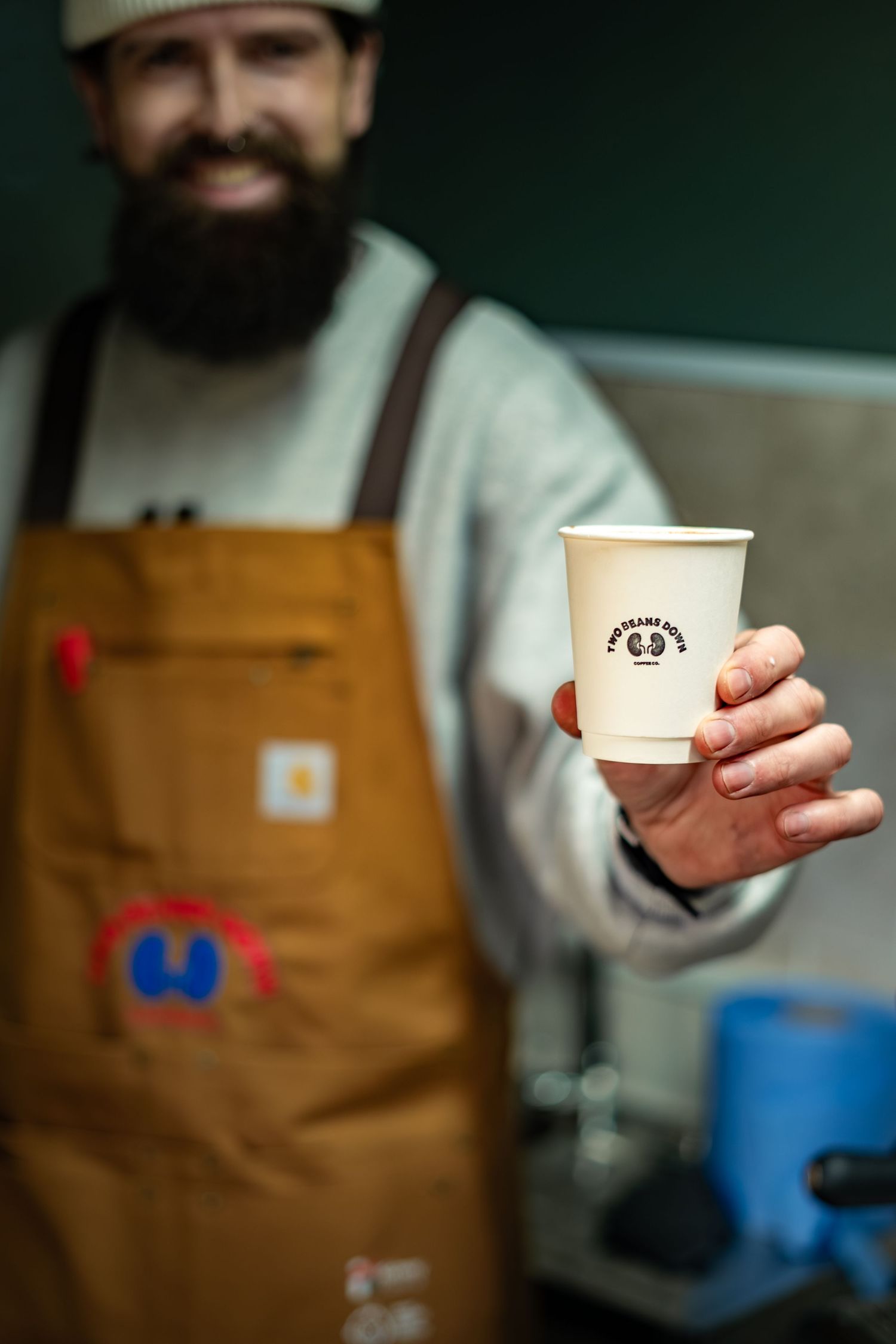 Smiling bearded barista extending a branded Two Beans Down paper coffee cup toward camera.