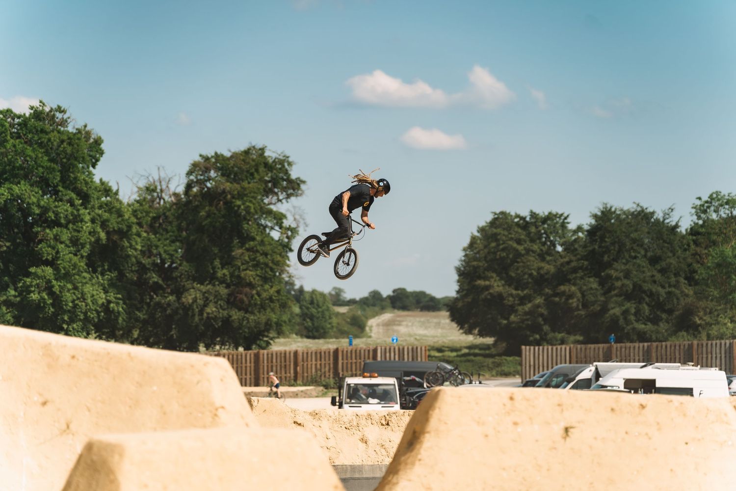 BMX cyclist performing a high aerial trick over sandy dirt jumps at an outdoor bike park on a sunny day.