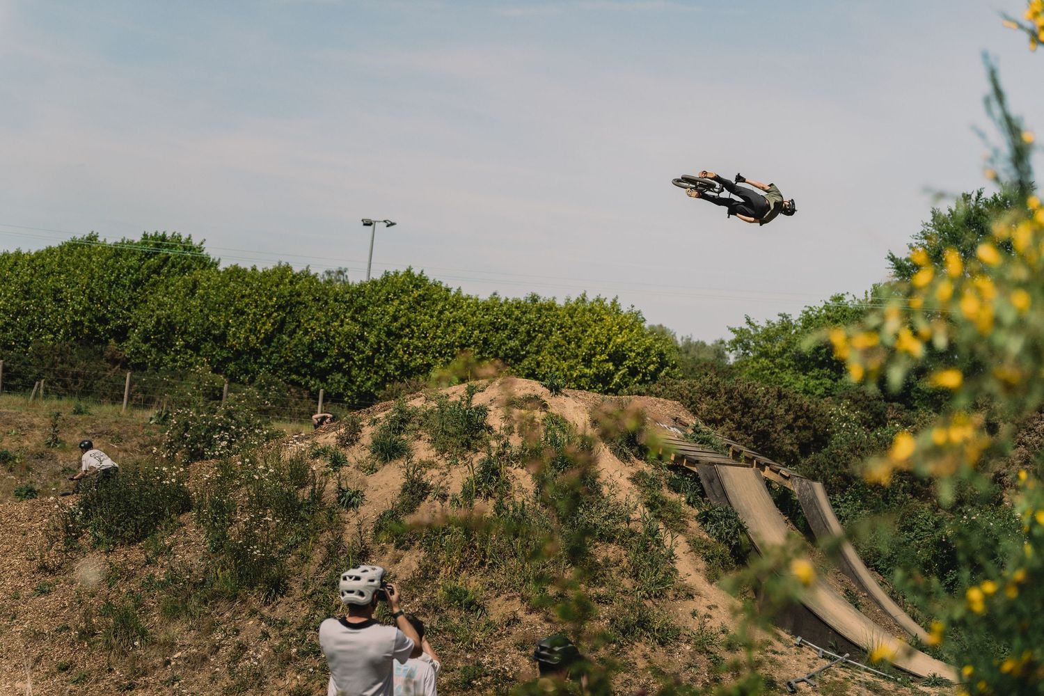 A BMX rider performs a high aerial trick over a dirt jump while a spectator watches below.