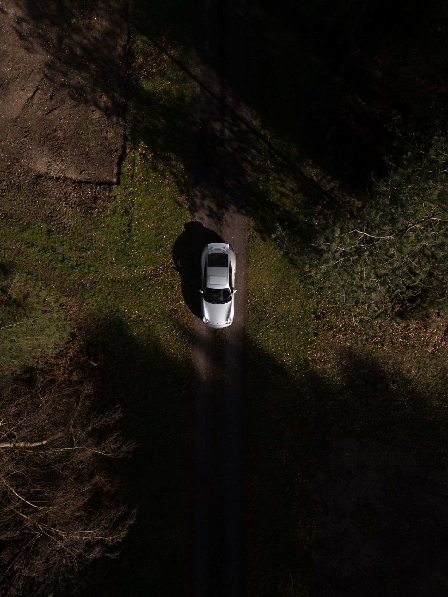 Bird's eye drone view of a silver Porsche 911 on a narrow dirt path between shadowed grass and woodland.
