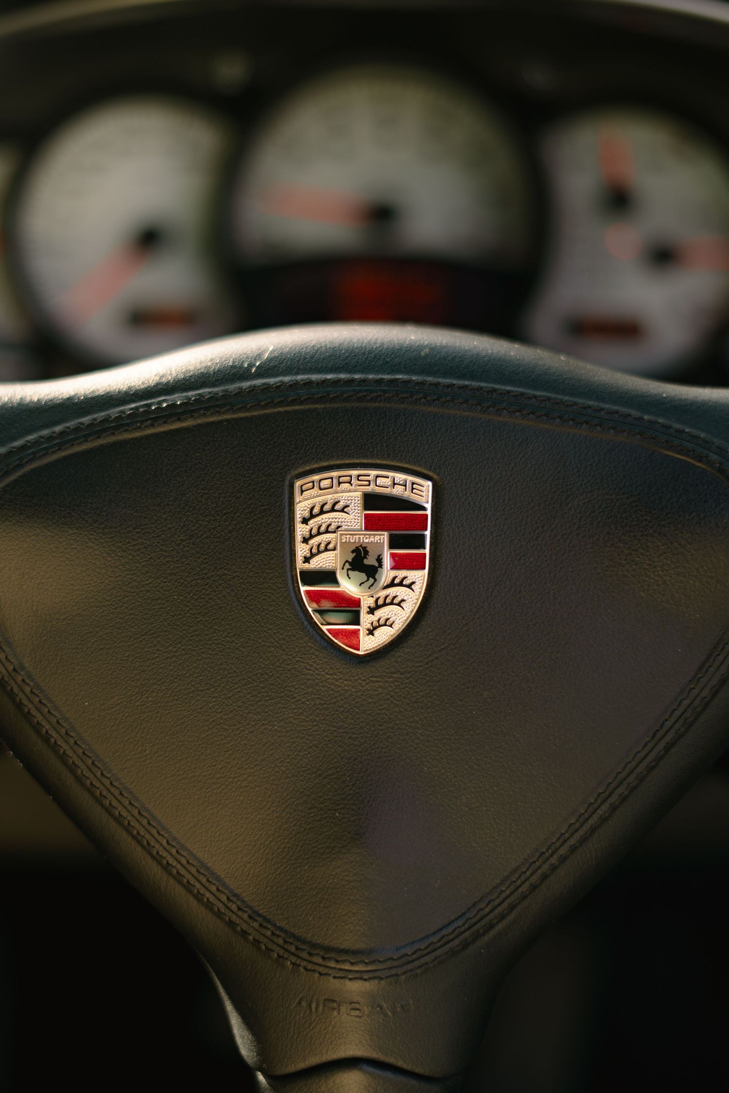 Close-up of Porsche crest logo on black leather steering wheel with gauges.