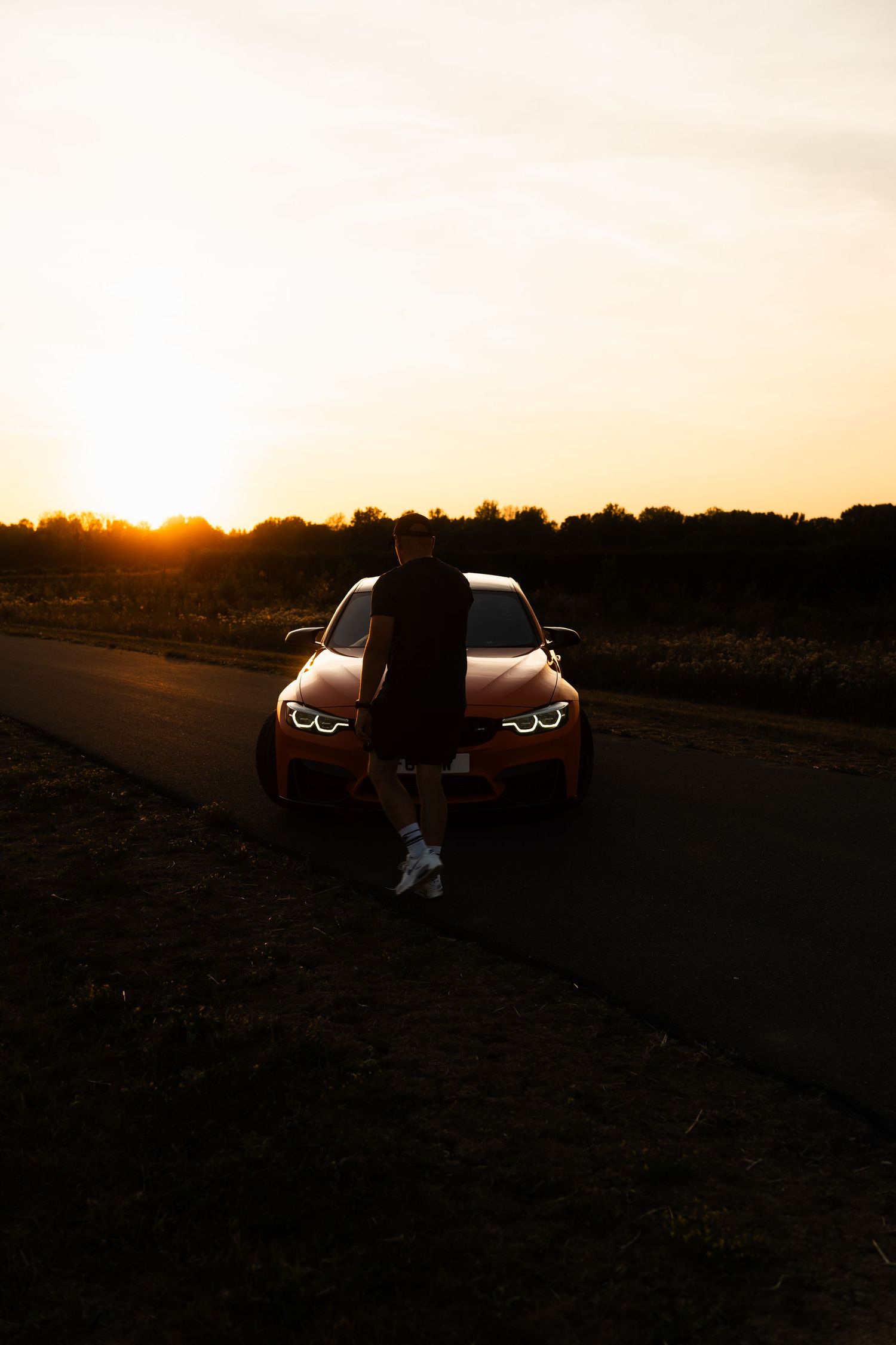 Person leaning on a red BMW car on a road at sunset with golden sky and silhouetted trees in background.
