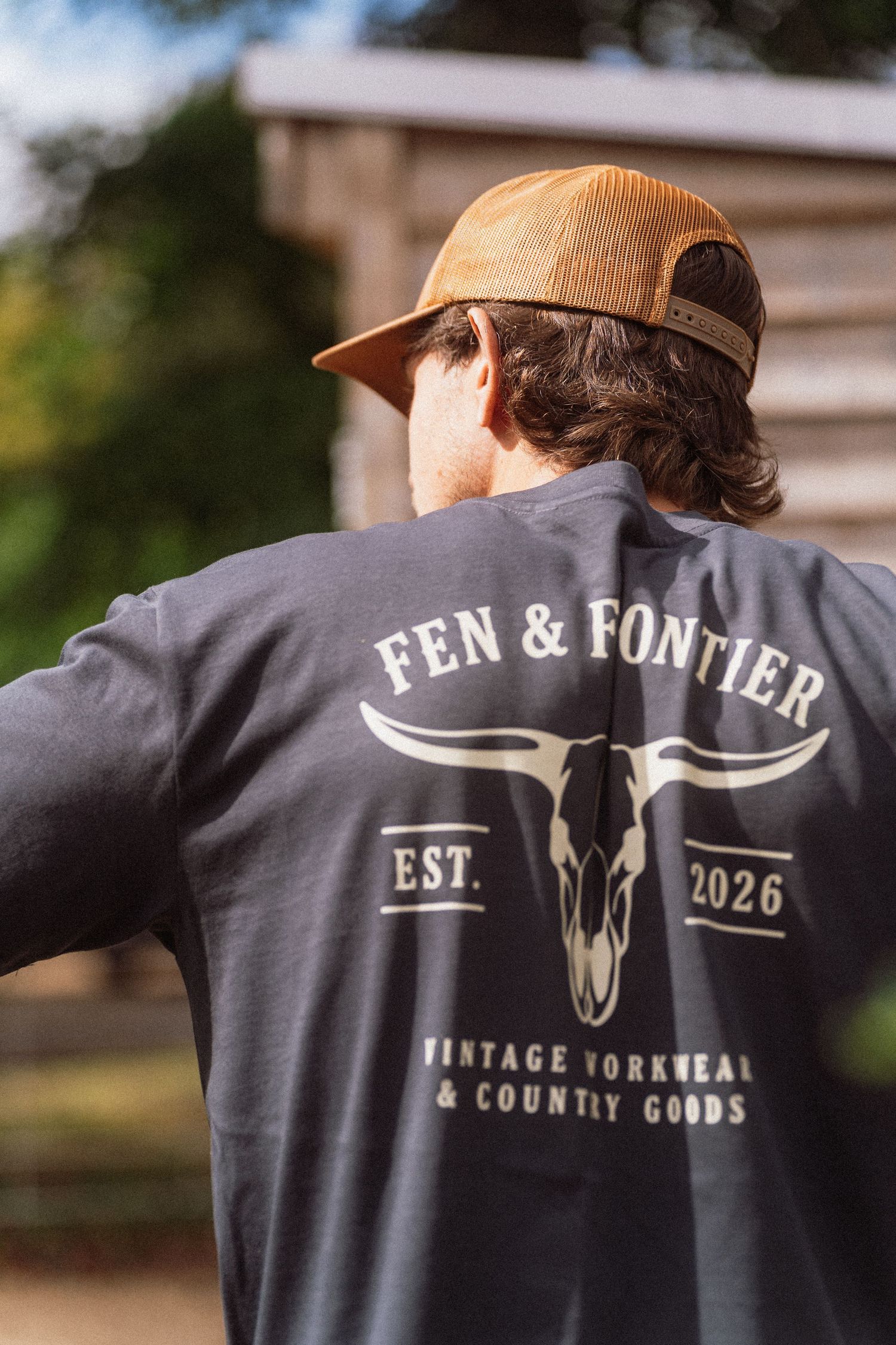 Man wearing a dark Fen & Fontier vintage workwear t-shirt and tan snapback cap, viewed from behind.