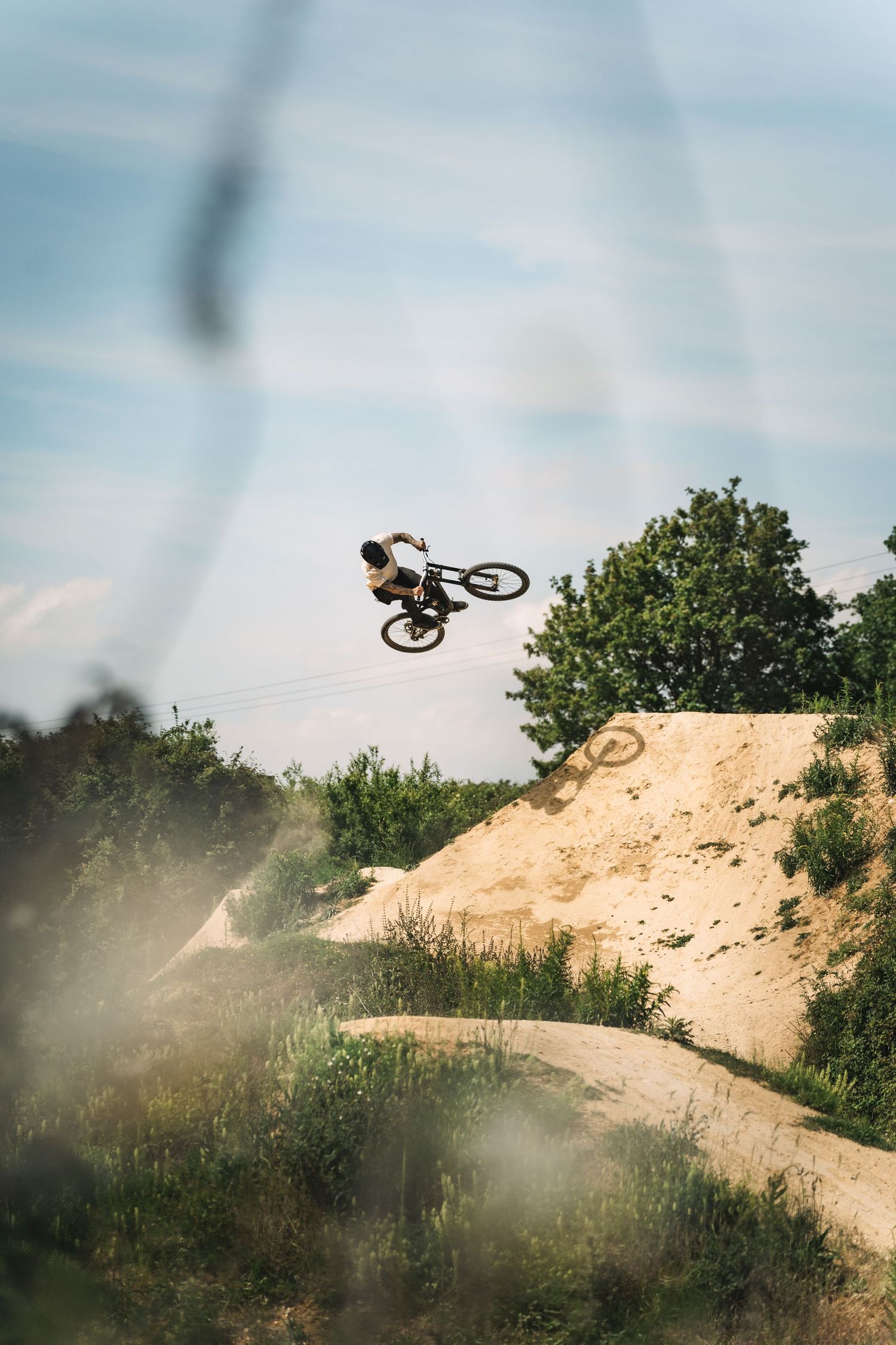 BMX rider performing a high aerial trick over sandy dirt jumps at an outdoor bike park with green trees in background.