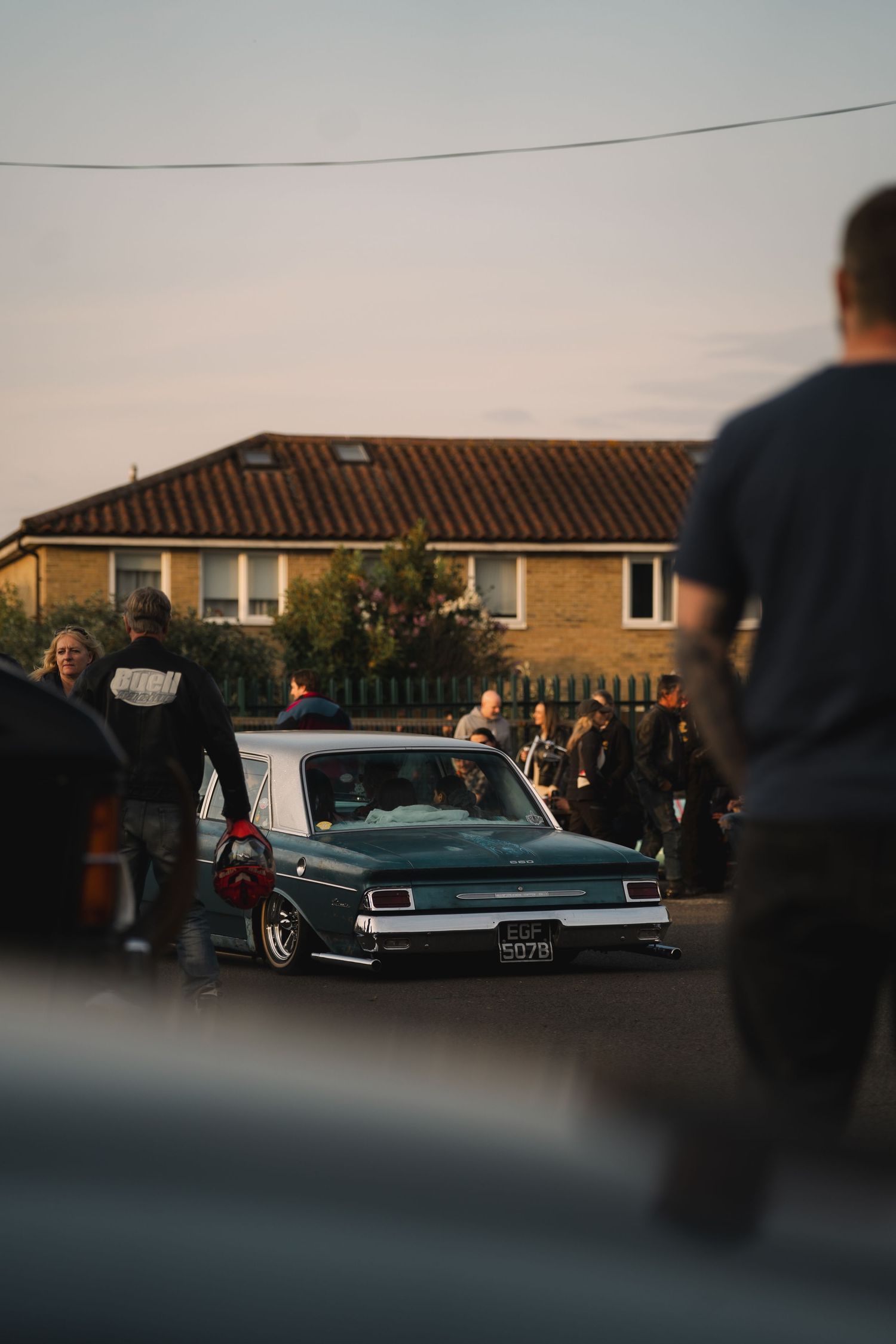 Lowered vintage teal sedan at a car meet surrounded by spectators near a brick building at dusk.