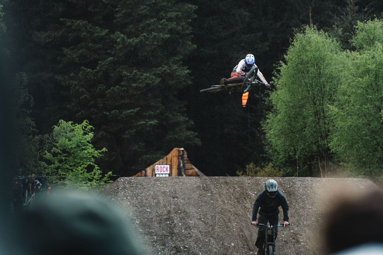 A mountain biker performs a high aerial jump over a dirt ramp at a bike park surrounded by dense green trees.