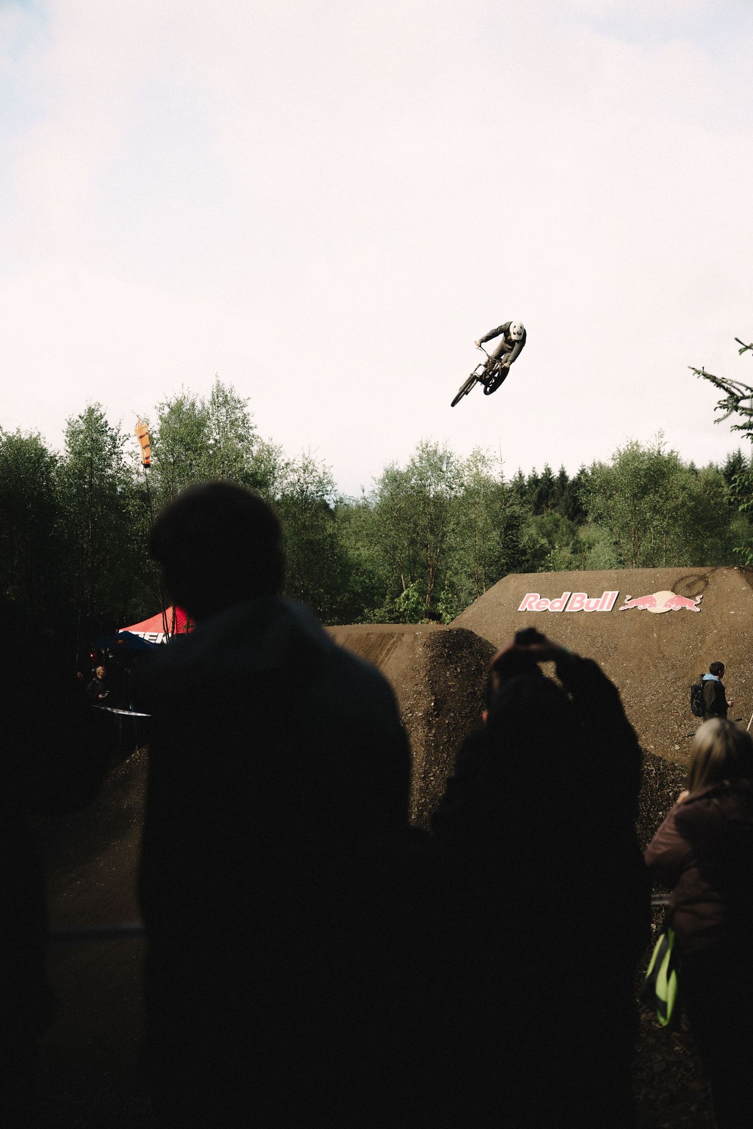 Motocross rider performs high aerial trick at Red Bull dirt jump event while silhouetted crowd watches below.