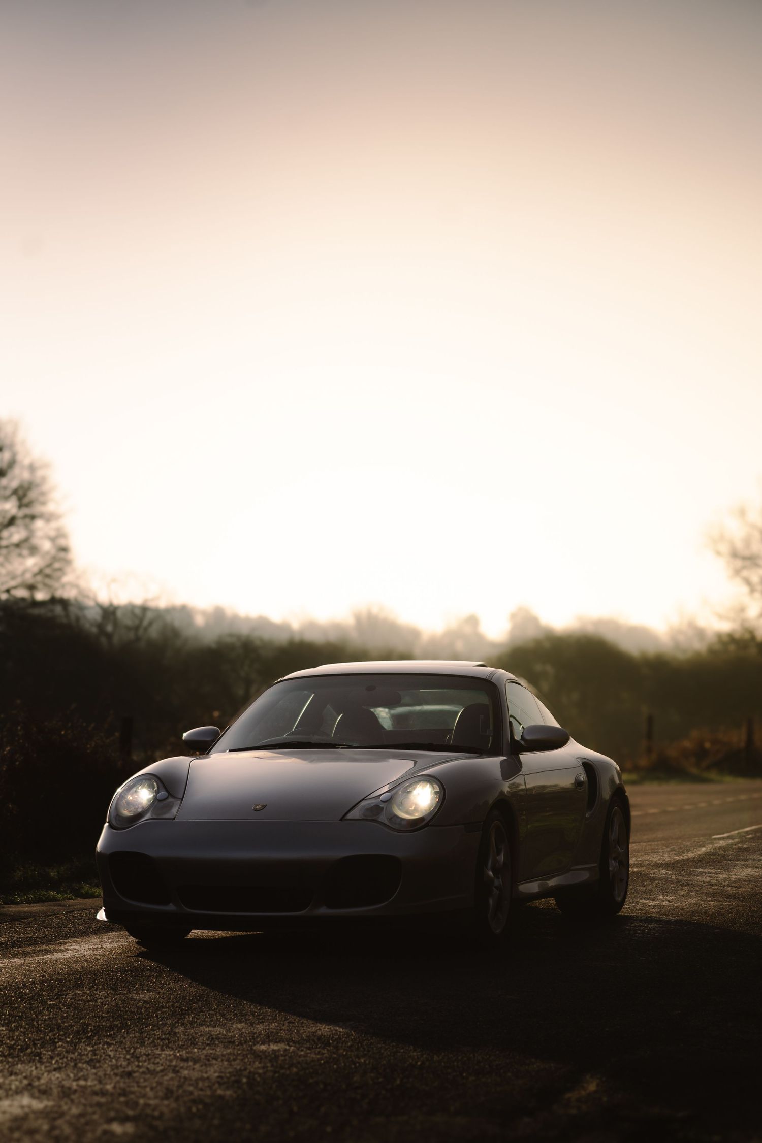 Silver Porsche 911 Turbo with headlights illuminated on a country road at golden hour sunset.
