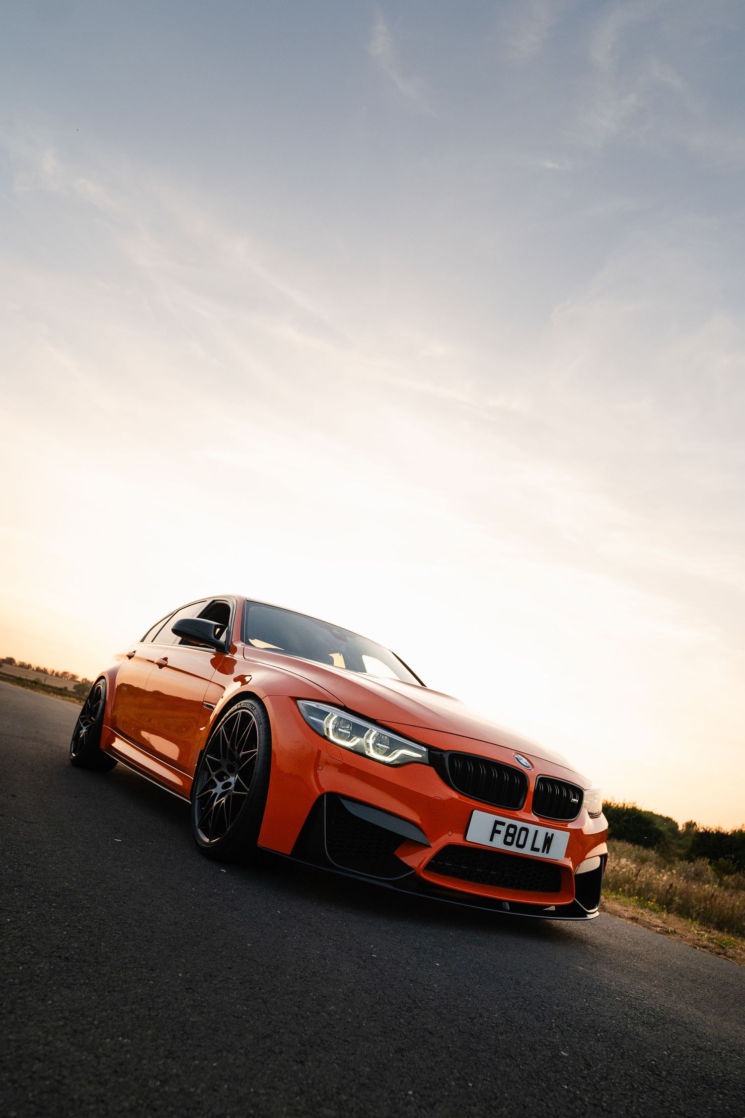 Orange BMW M3 F80 on a road at sunset, featuring black wheels and carbon fiber accents, with a dramatic sky backdrop.