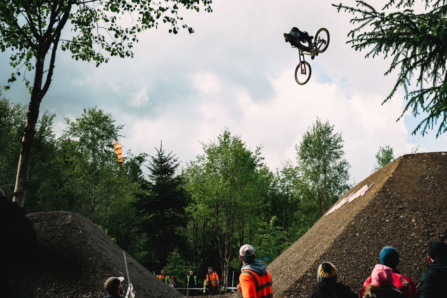 A mountain biker performs a high aerial trick over dirt jumps surrounded by trees with spectators watching below.