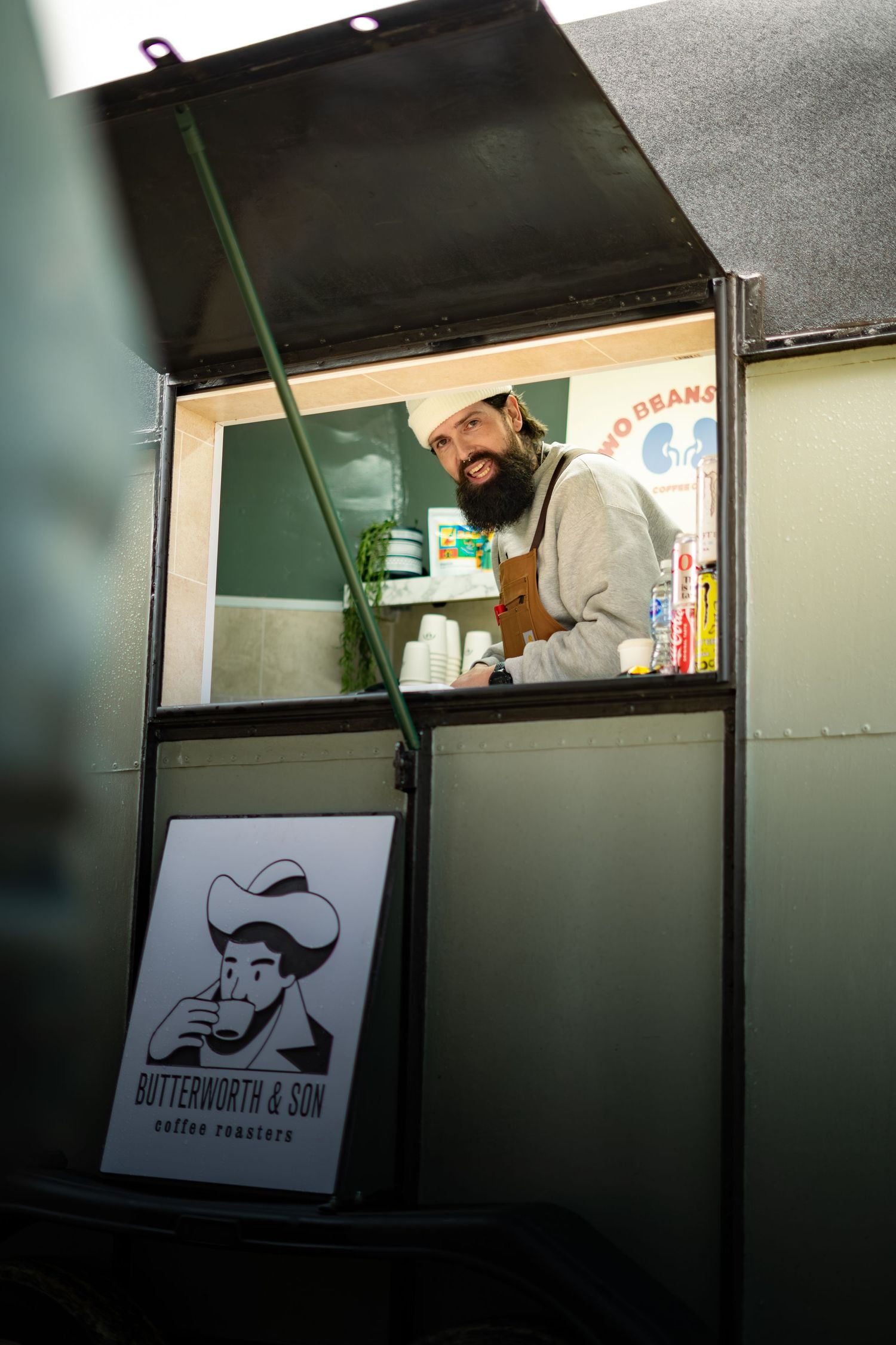 Smiling bearded barista stands in Butterworth & Son coffee truck window wearing brown apron.