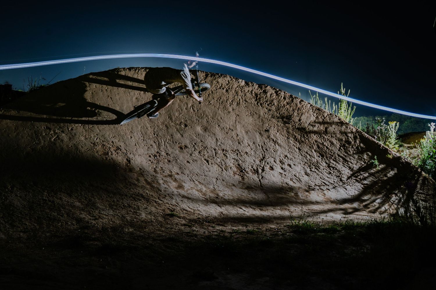 Mountain biker performing a nighttime jump on a dirt trail, illuminated by light trails against a dark sky.