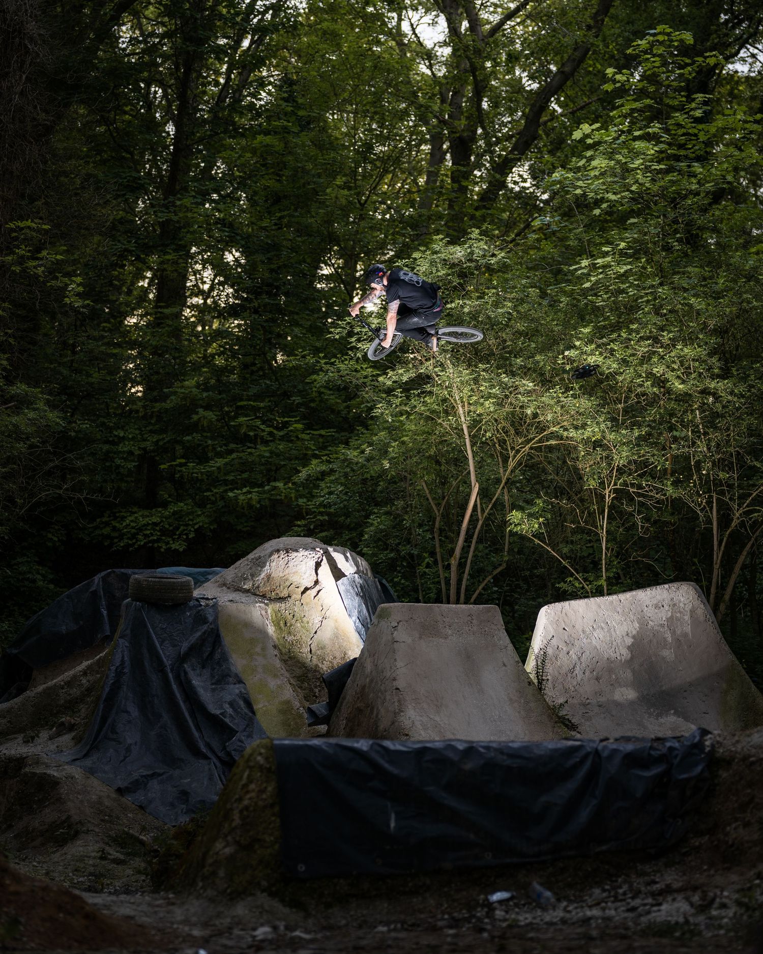 BMX rider performing a high aerial trick over concrete dirt jumps in a forest setting.