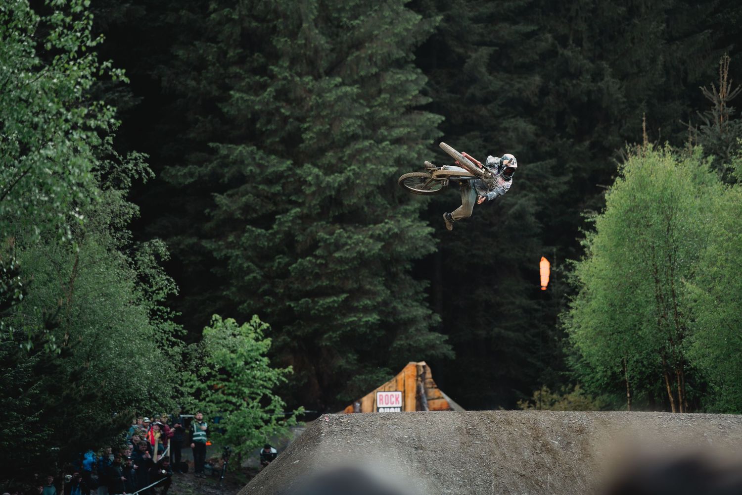 Motocross rider performing a high aerial trick off a dirt ramp surrounded by dense green forest trees.