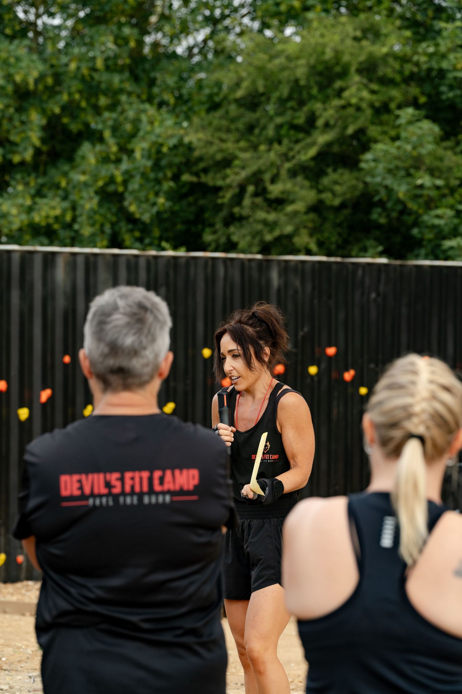 A fitness instructor leads an outdoor Devil's Fit Camp workout session, holding exercise equipment while participants follow along.