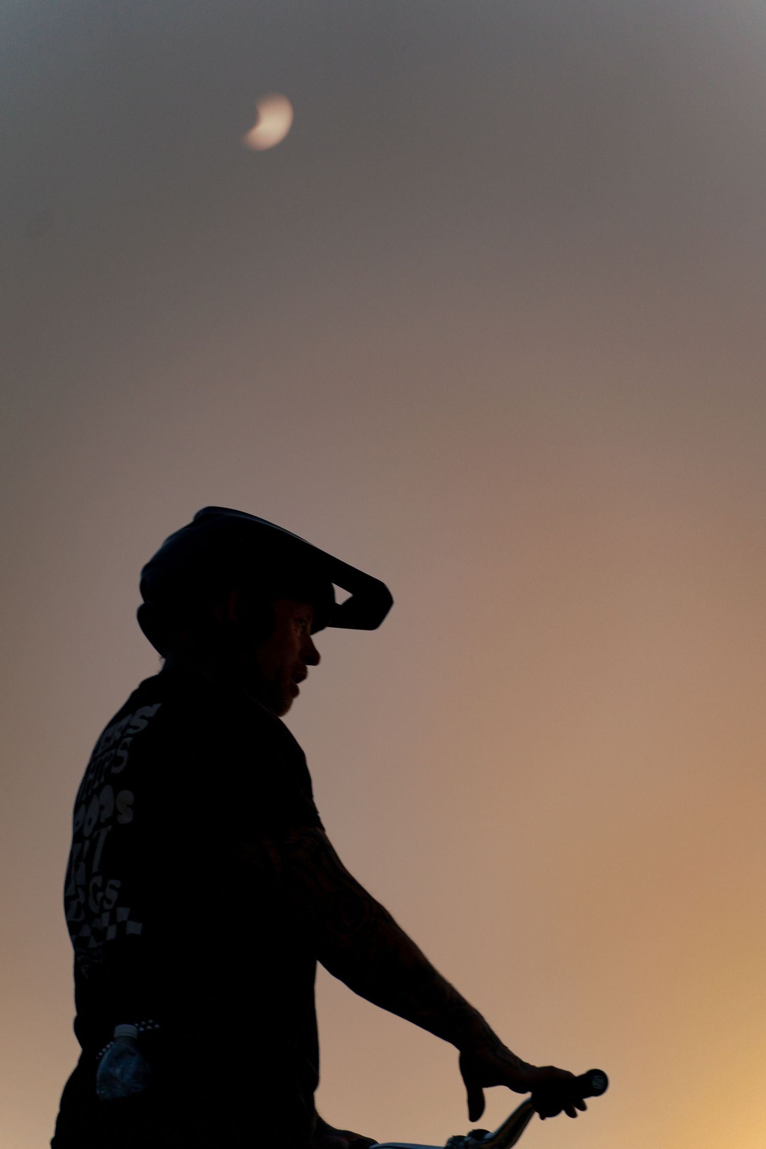 Silhouette of a cyclist wearing a helmet against a twilight sky with a crescent moon above.