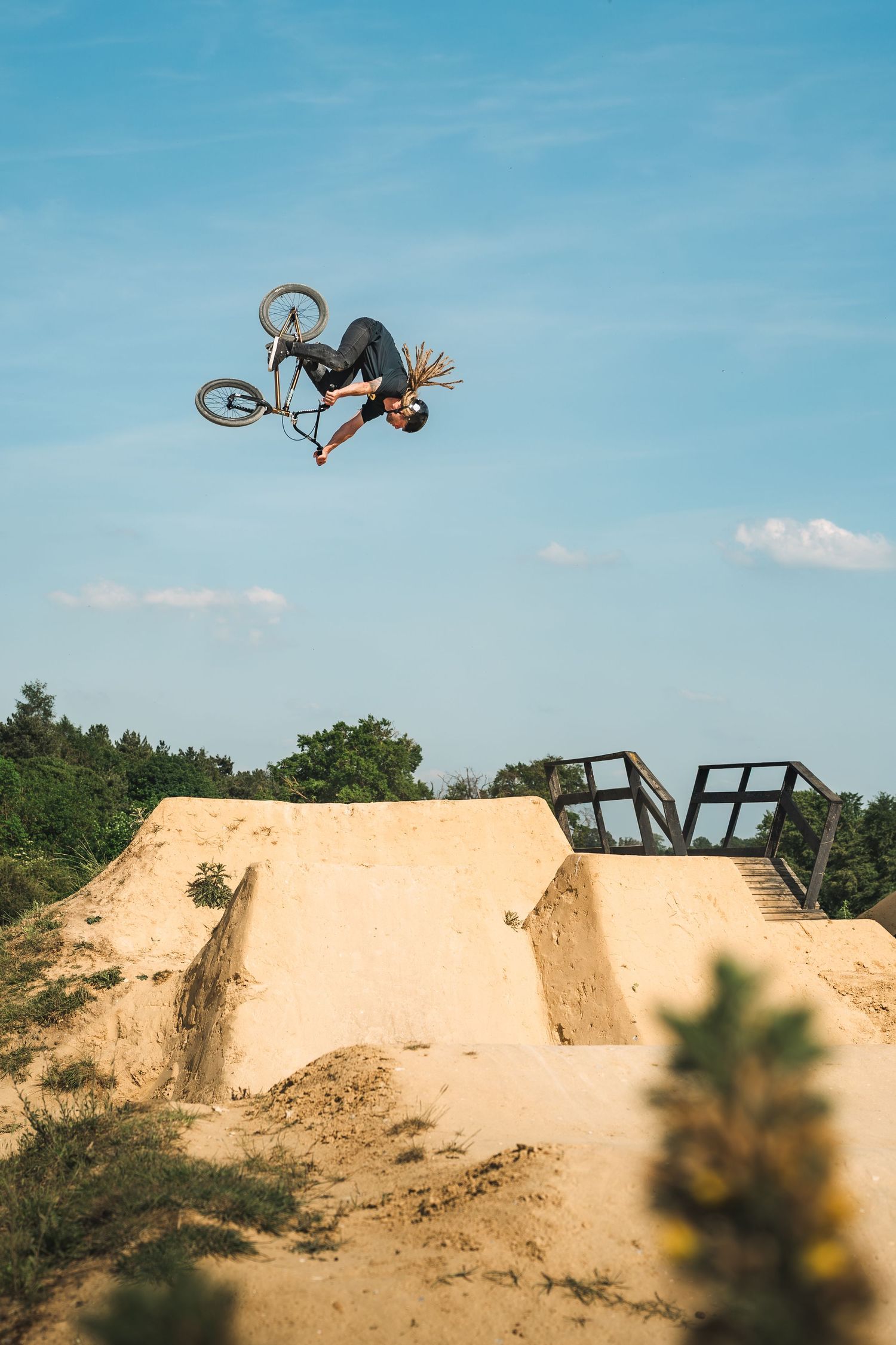 BMX rider performing an inverted aerial trick above sandy dirt jumps at an outdoor bike park.