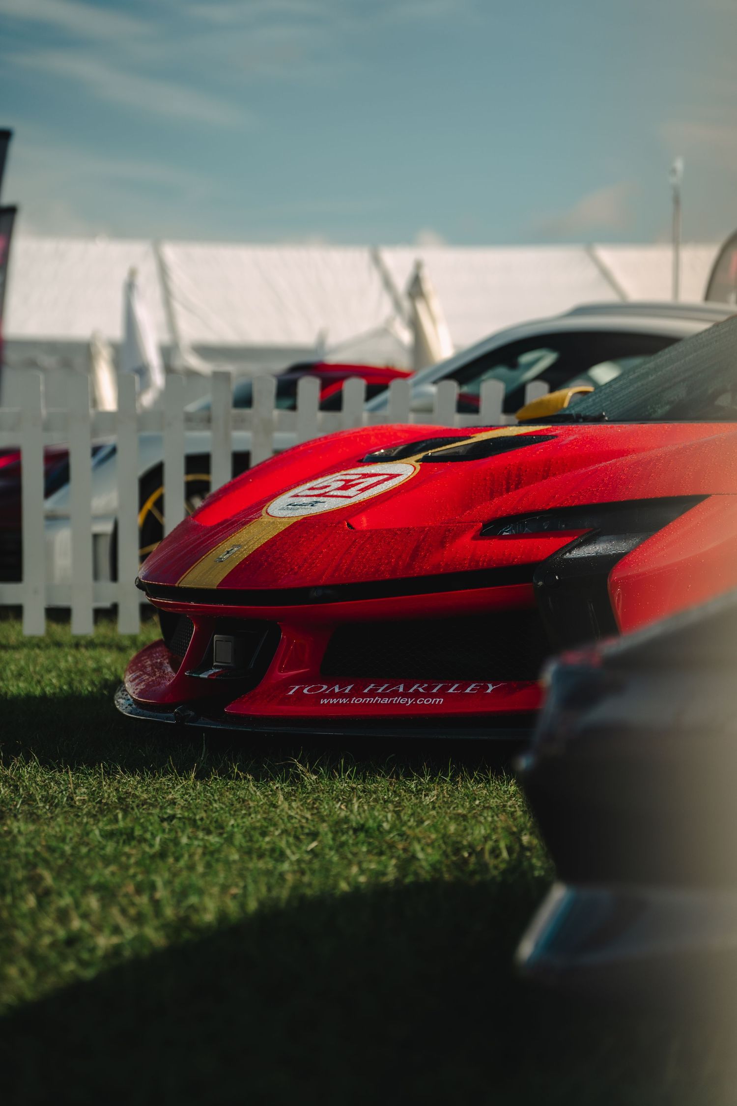 Red Ferrari with gold stripes and Tom Hartley branding on display at an outdoor motorsport event on green grass.
