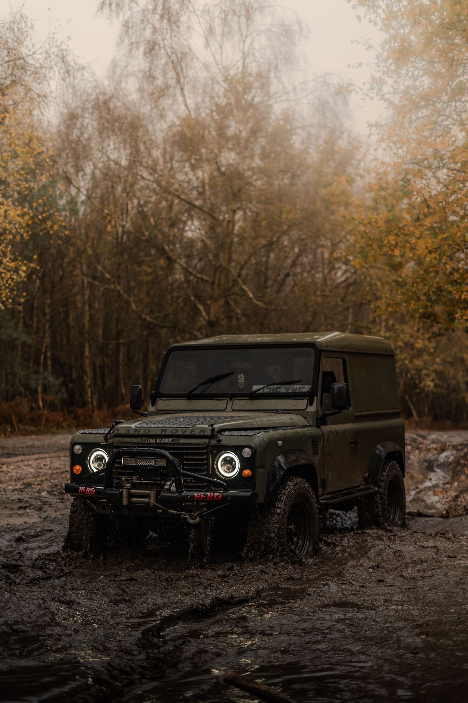 Modified Land Rover Defender with halo headlights navigating a muddy off-road trail in an autumn forest.