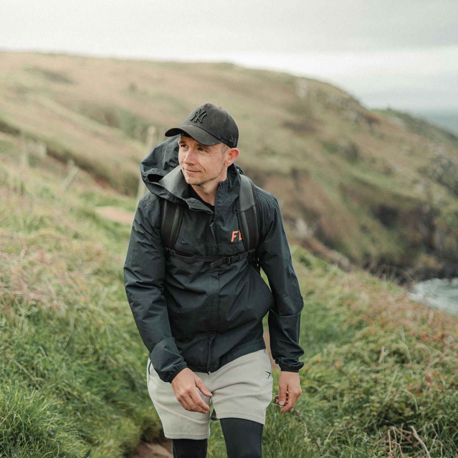 Male hiker wearing black jacket and cap with backpack on scenic coastal cliff trail.