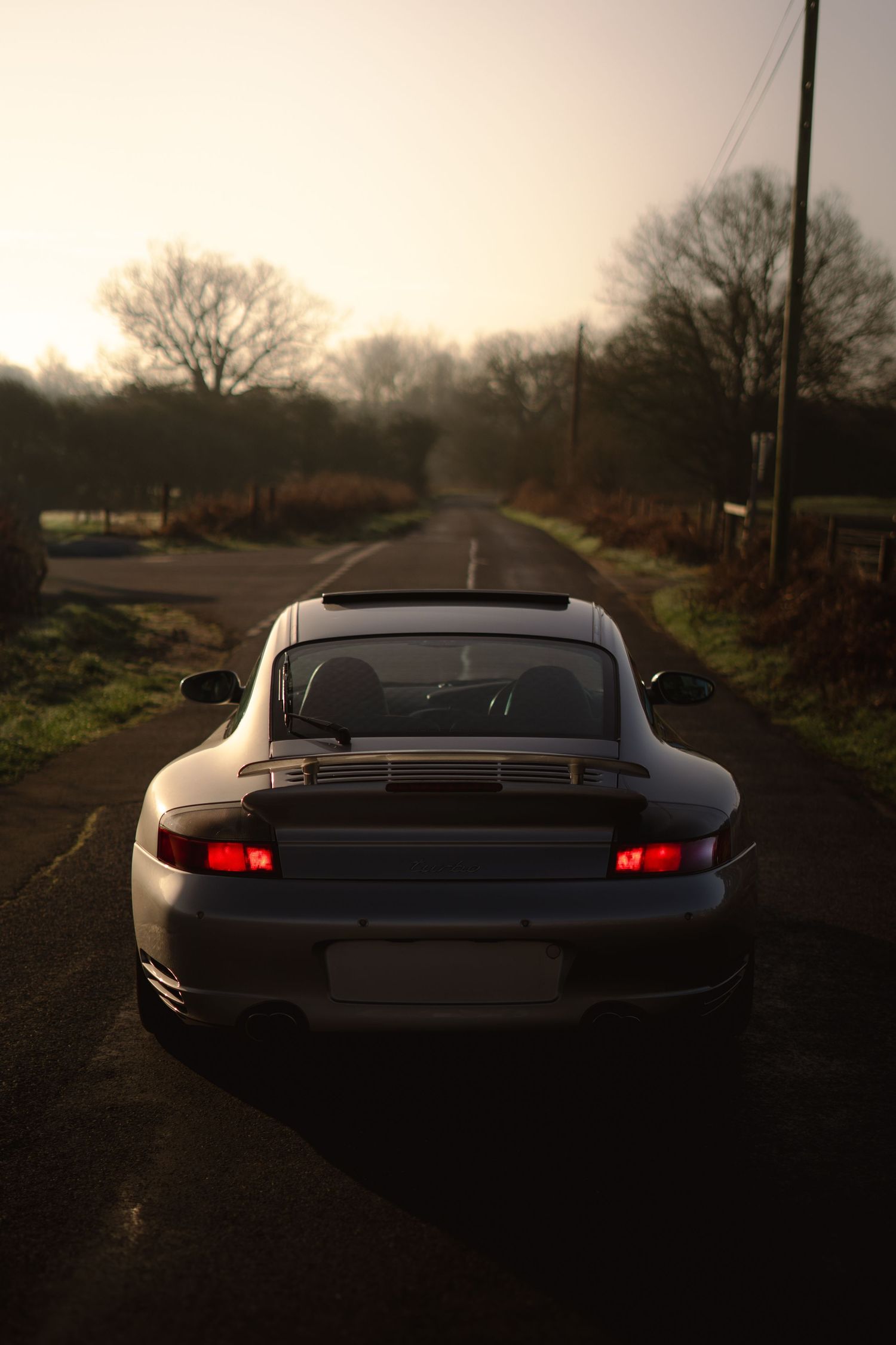 Silver Porsche 911 rear view on a rural road at golden hour with glowing red tail lights and misty sky.