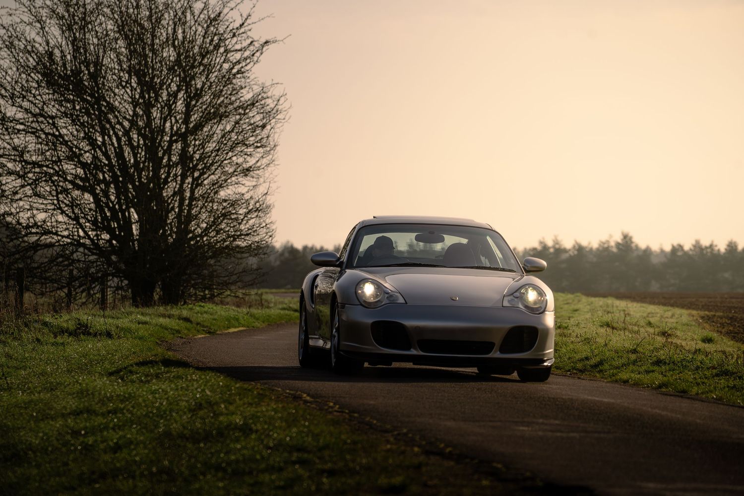 Silver Porsche 911 Turbo on a country road at dusk with glowing headlights and misty sky.