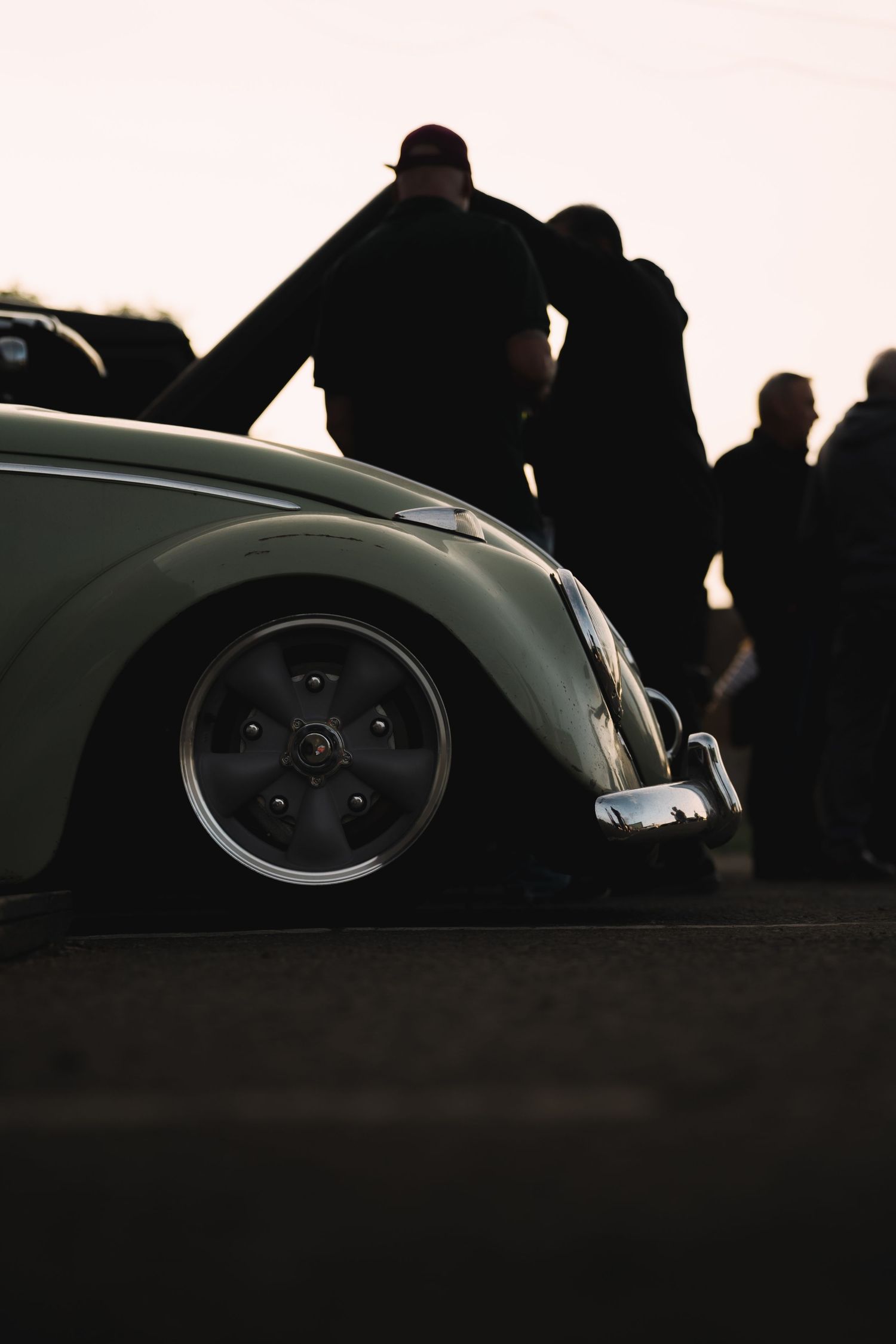 Low-angle view of a classic VW Beetle with custom wheels, silhouetted figures gathered around at dusk.
