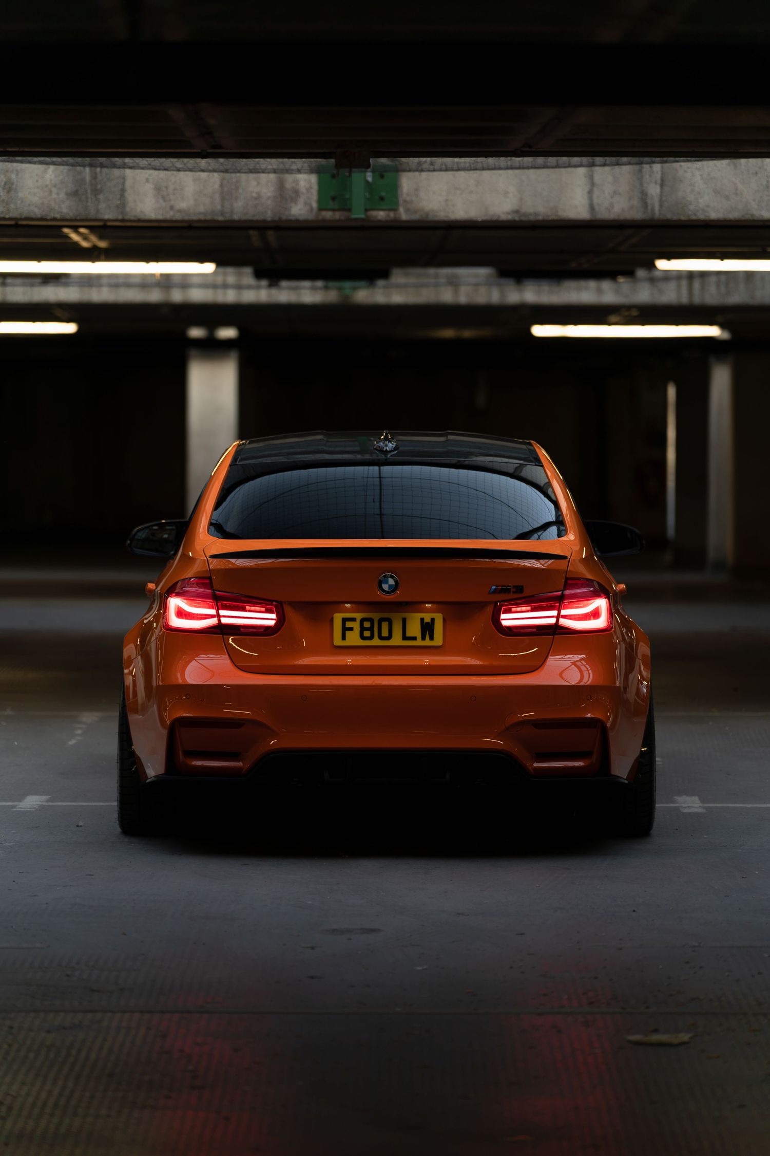 Orange BMW M3 F80 rear view in parking garage with glowing taillights and UK license plate F80 LW.
