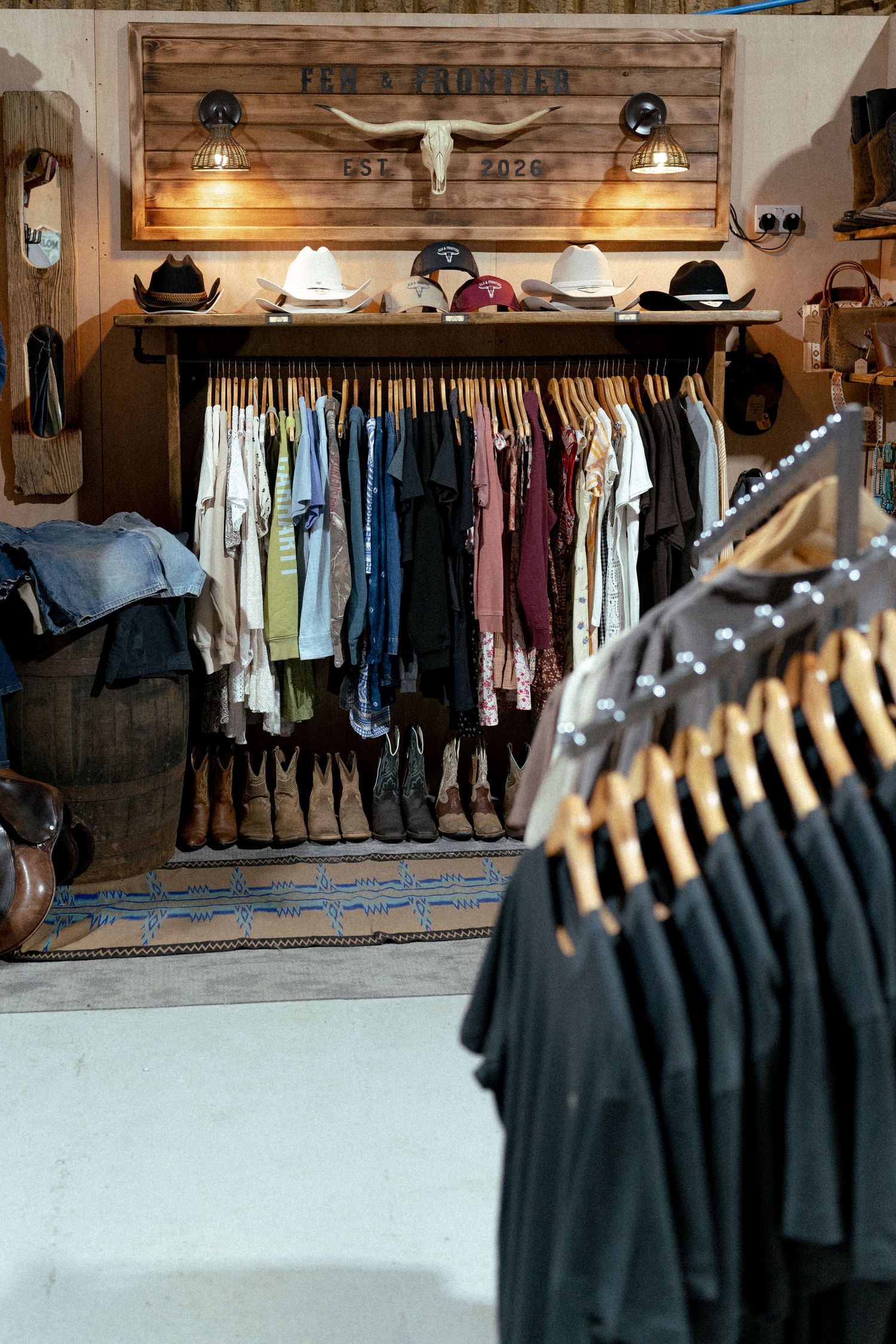 Western boutique store interior with cowboy hats, boots, and colorful clothing on rustic wood racks beneath bull skull decor.