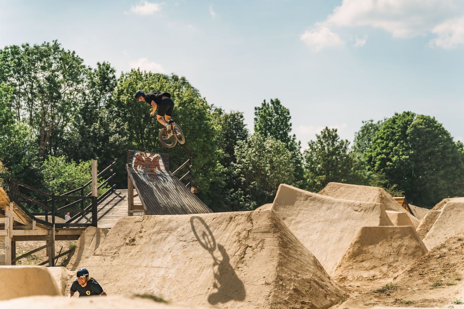 BMX rider performing a high jump trick over dirt mounds at an outdoor bike park on a sunny day.