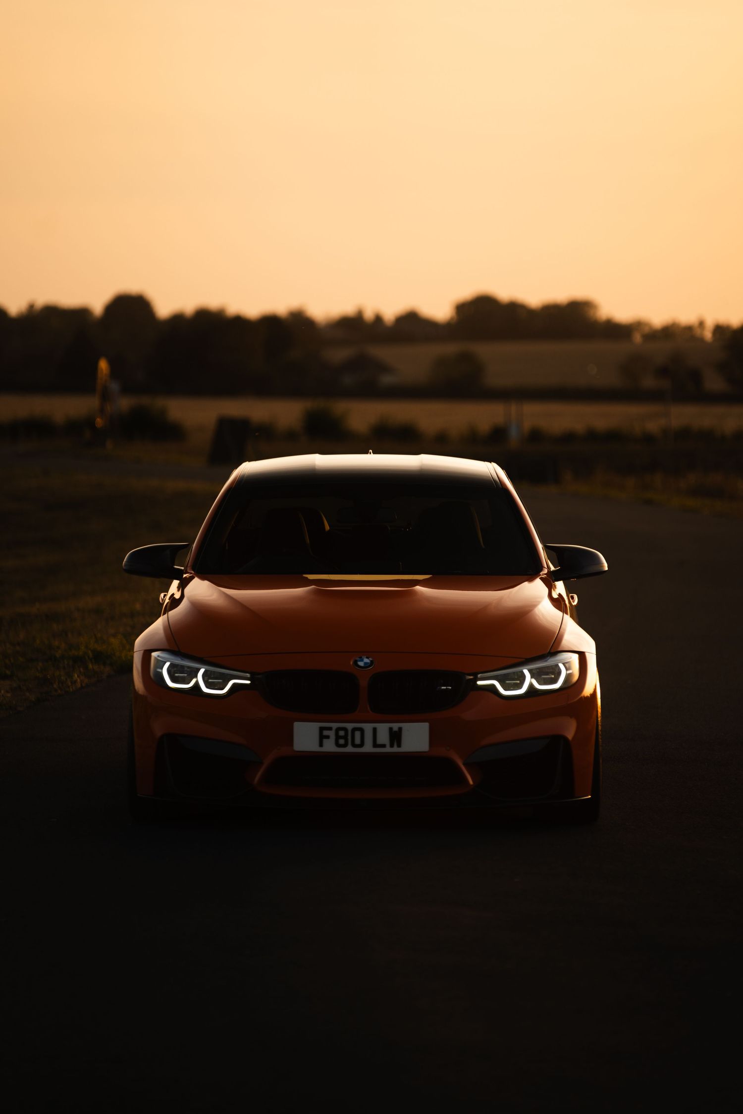 Orange BMW M3 F80 photographed from the front at golden hour sunset on a rural road.