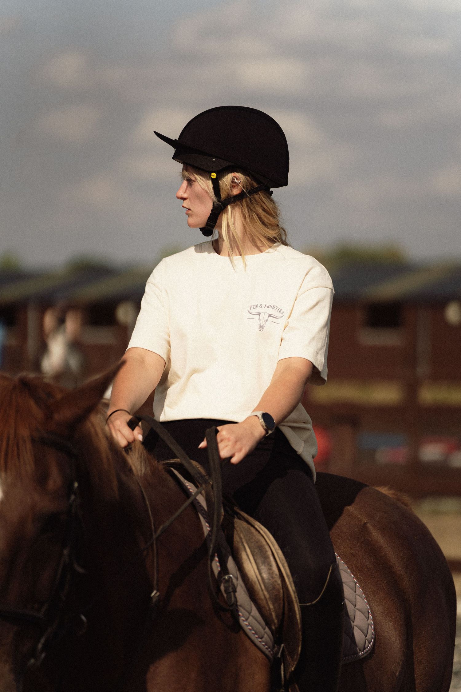 Young blonde woman in black helmet and white shirt riding a chestnut horse at an equestrian facility.