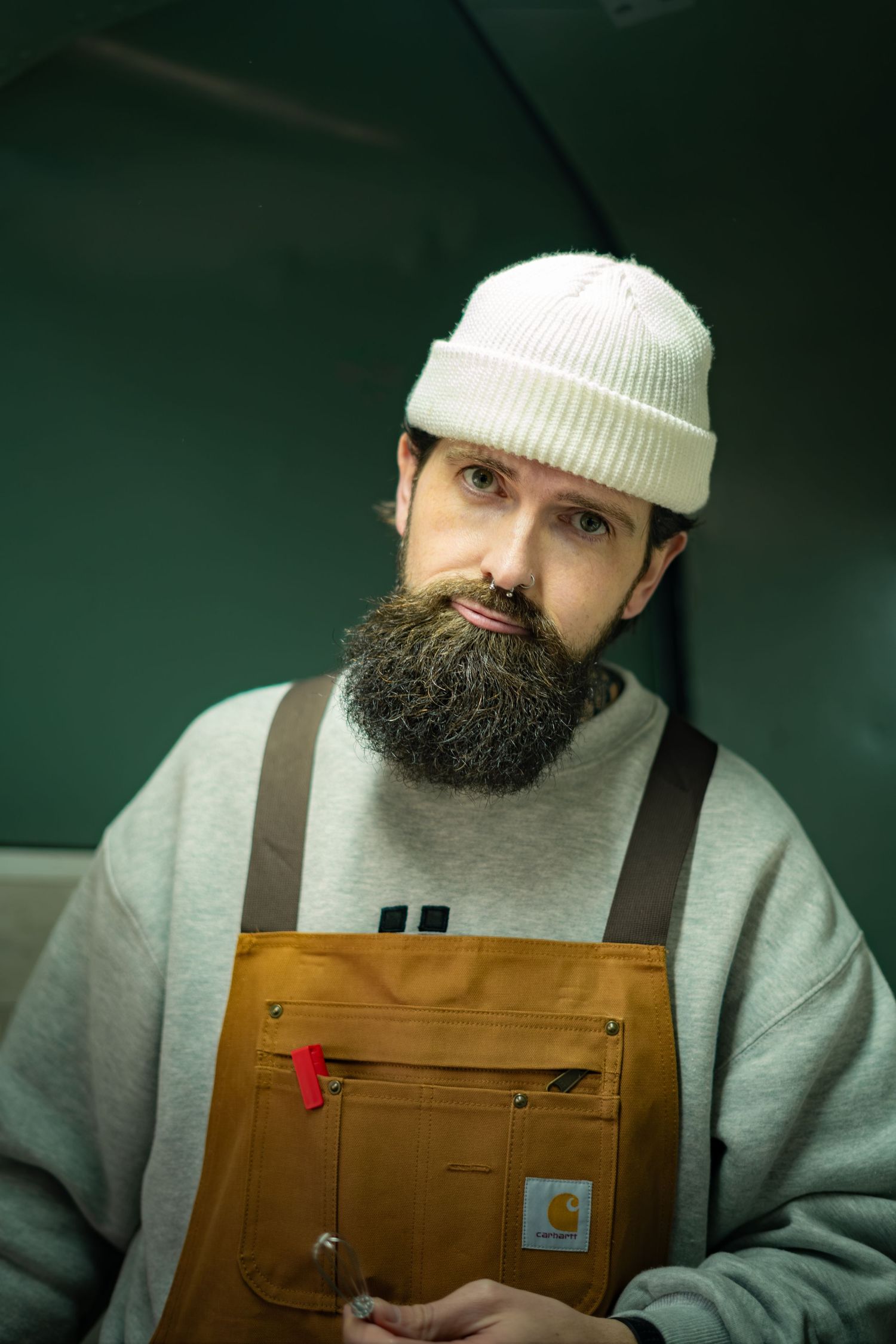 Bearded barista in white knit beanie and Carhartt brown overalls poses inside coffee truck.