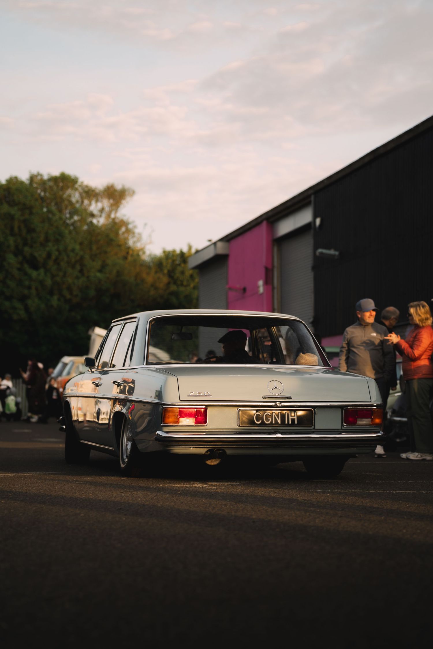 Vintage Mercedes-Benz sedan with license plate CGN1H parked at dusk near a black building with onlookers.